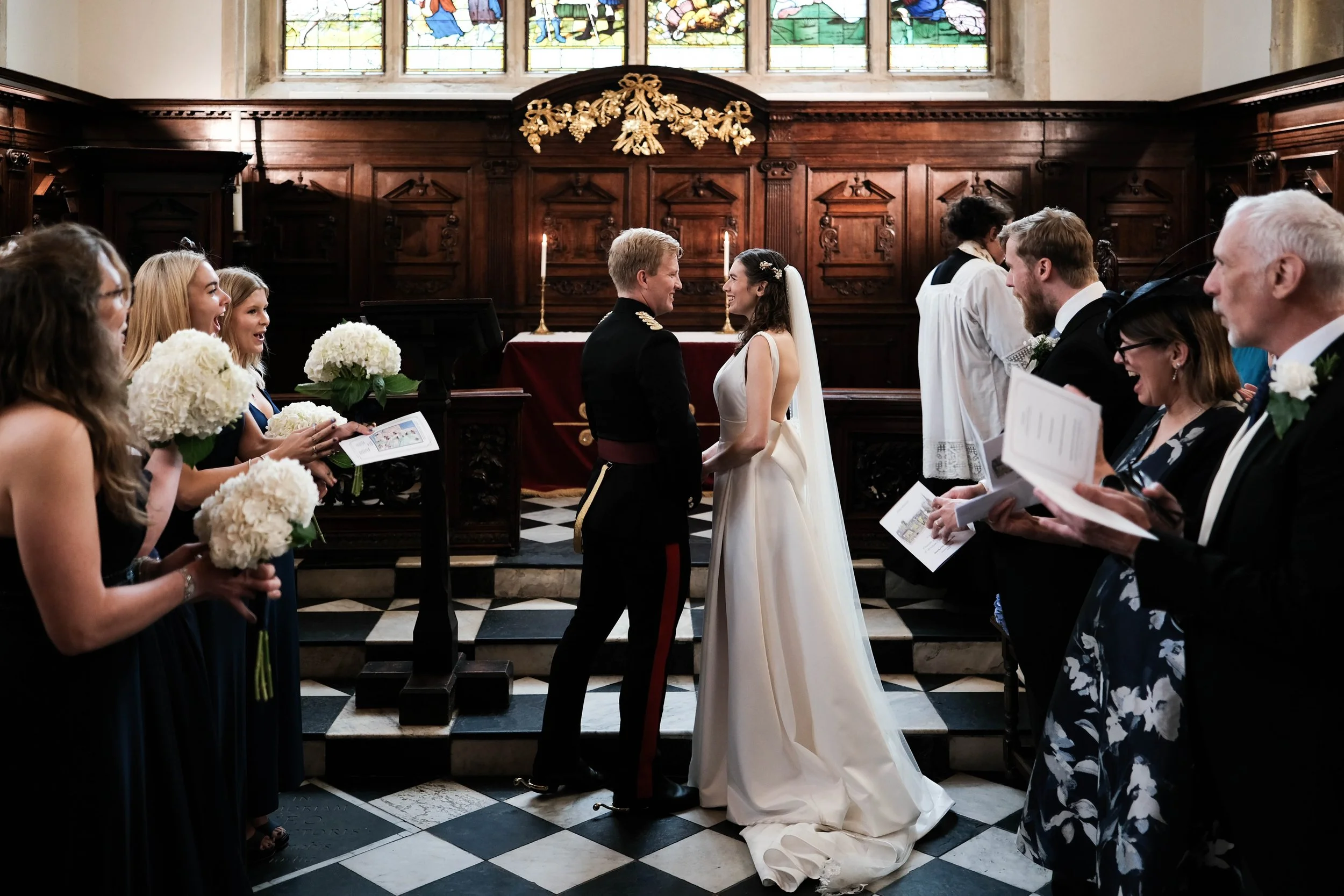 A wedding ceremony in a church with the bride and groom standing in front of the officiant, holding hands. The bride is wearing a white gown with a veil, and the groom is in a military uniform. Bridesmaids on the left hold white bouquets, and groomsm