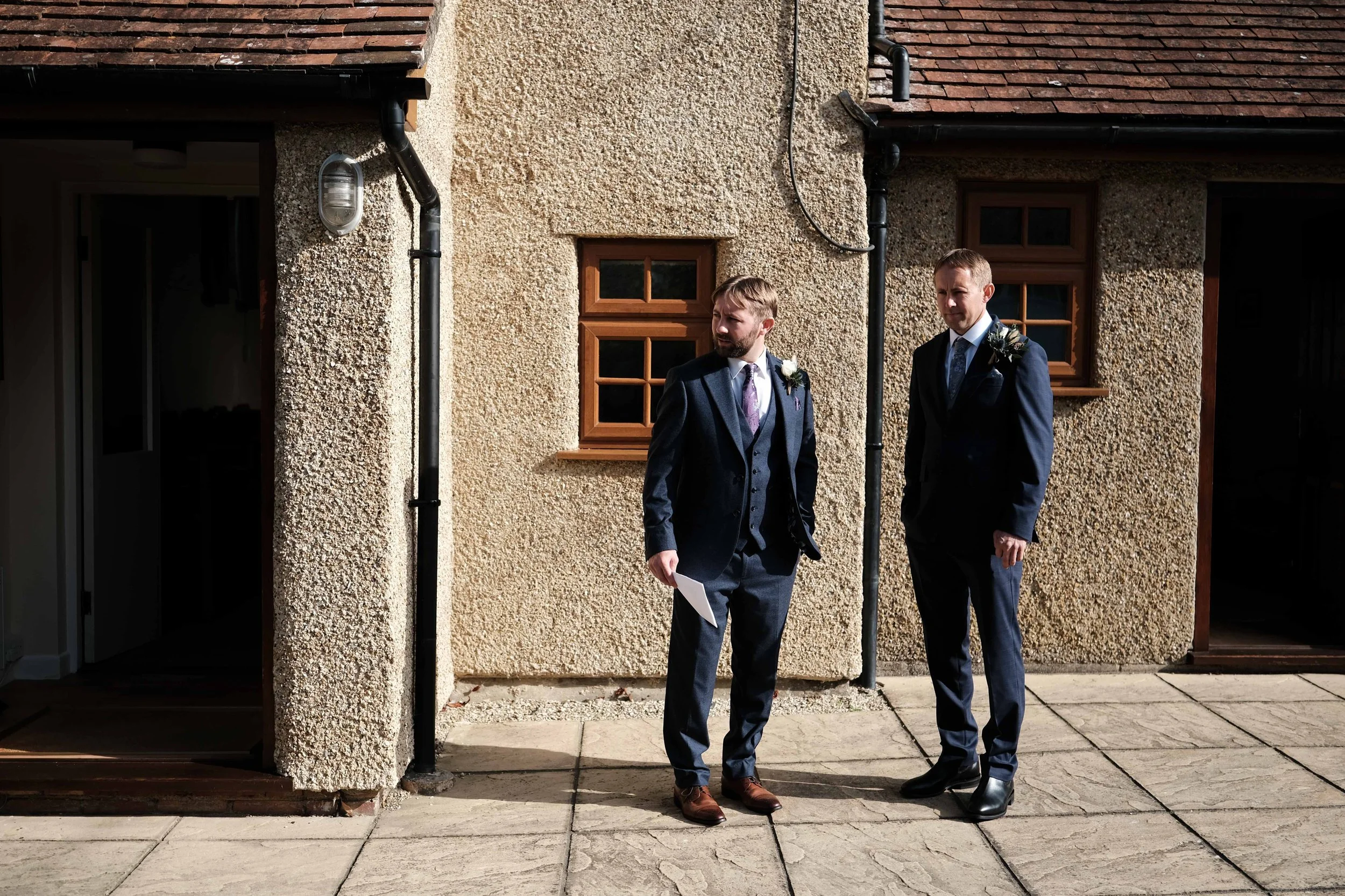 Two men in suits standing outside a house, one holding a paper, with sunlight casting shadows on the wall.
