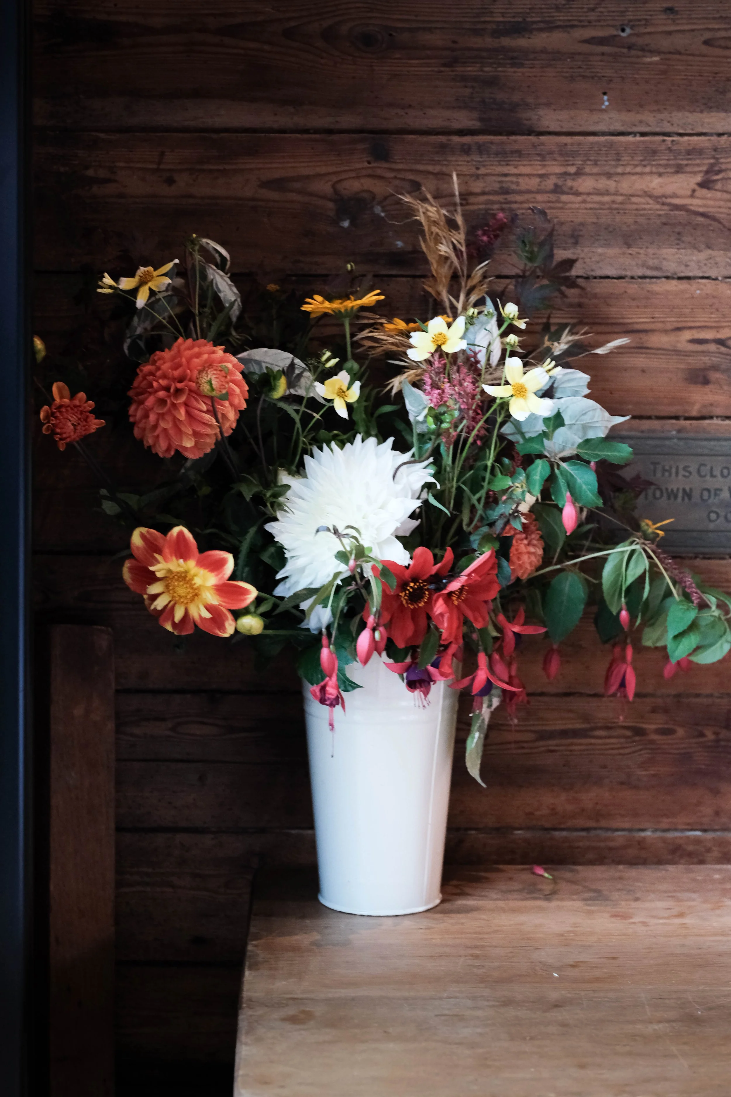 A bouquet of multicolored flowers in a white vase on a wooden surface against a wooden wall background.