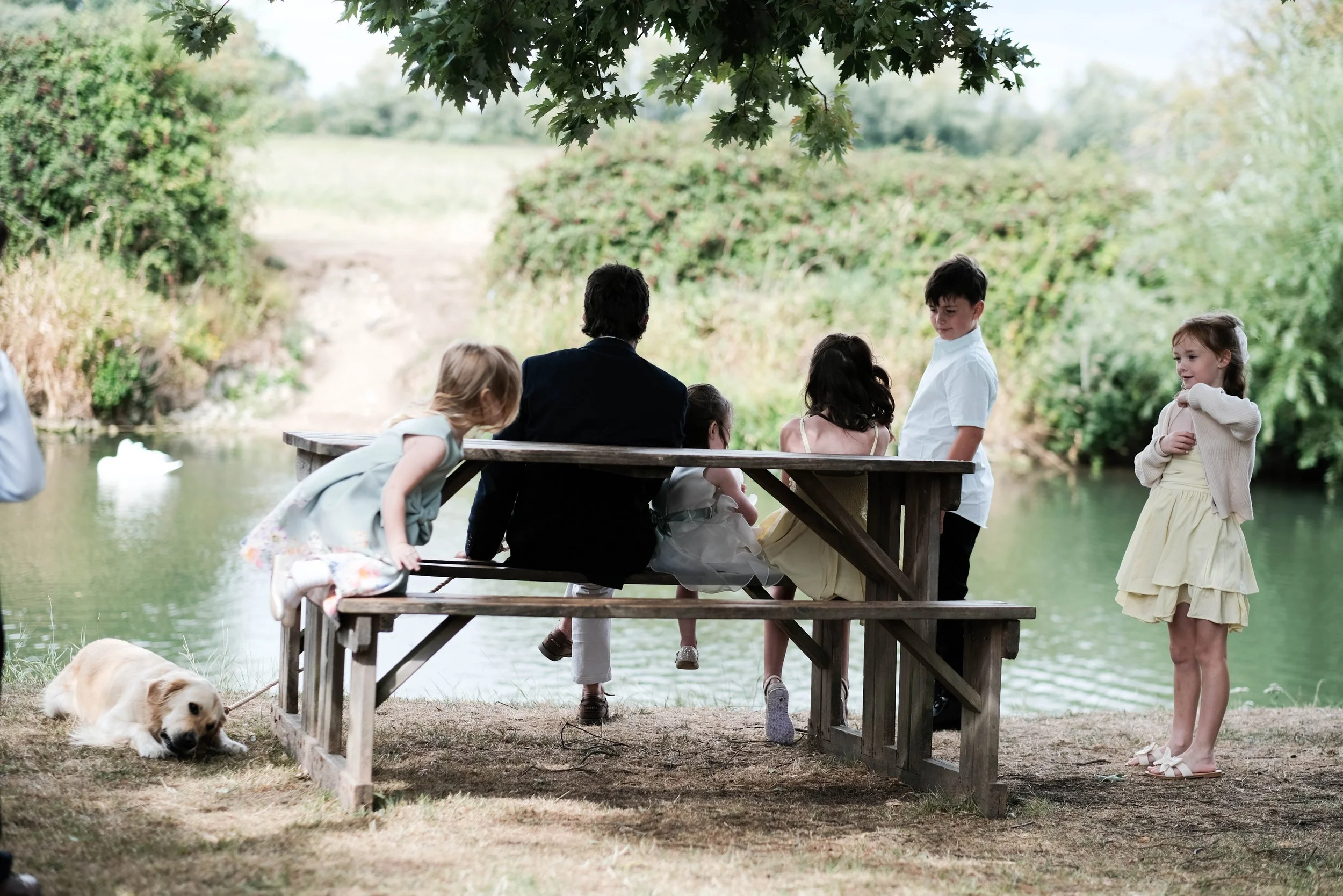 Children and an adult sitting on a picnic table by the water, with a dog lying on the ground nearby, under a tree with lush greenery in the background.