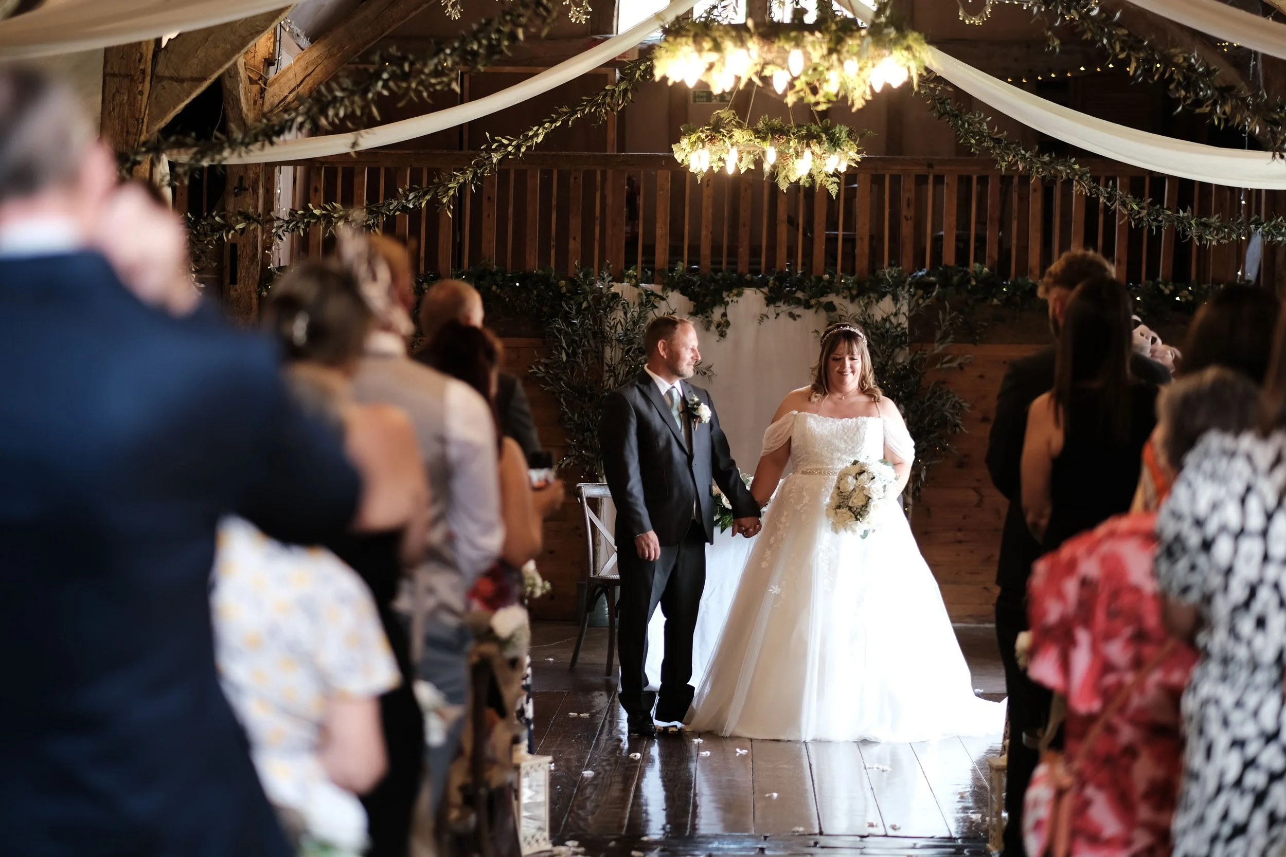 Wedding ceremony with bride and groom holding hands, surrounded by guests in a rustic decorated venue with wooden beams, greenery, and hanging lights.