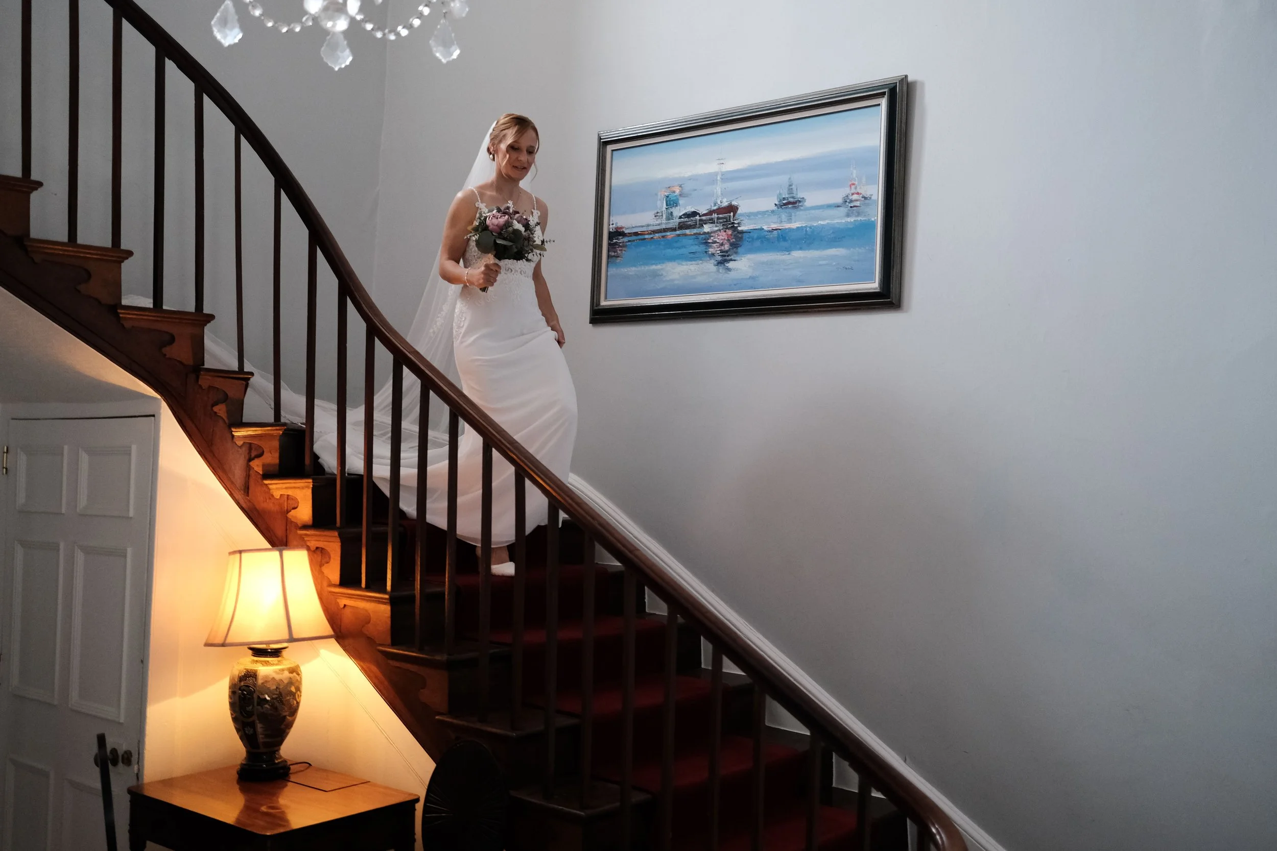A bride in a white wedding dress holding a bouquet of flowers walking down wooden stairs inside a house.