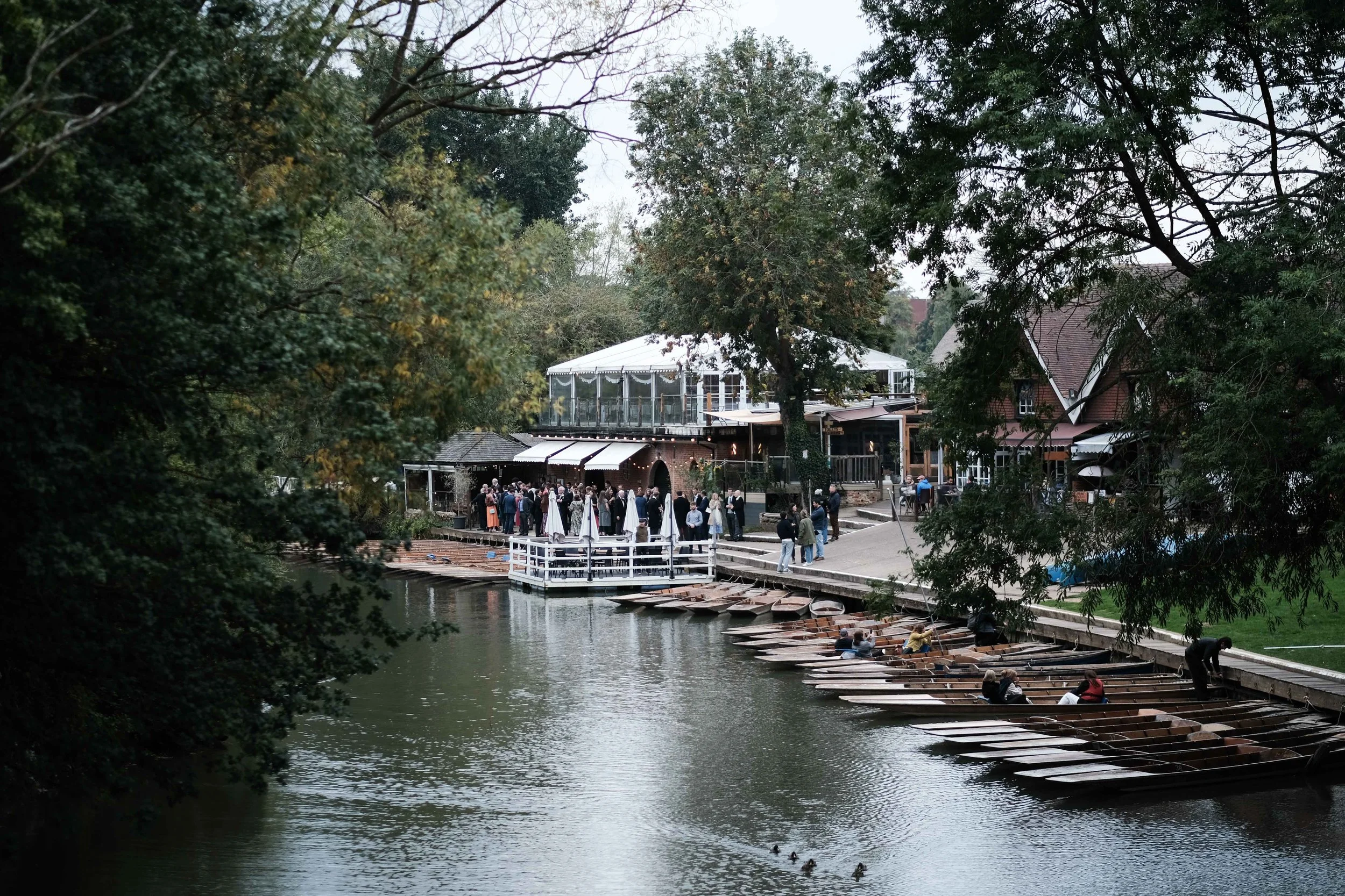 People gathered on a dock and boats in a river near a restaurant or event space surrounded by trees.