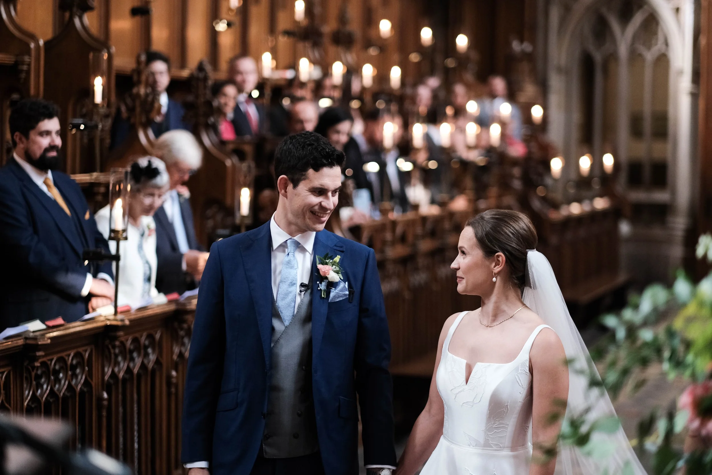 A bride and groom standing at the altar, holding hands and smiling at each other, with wedding guests seated in the background inside a church decorated with candles and flowers.