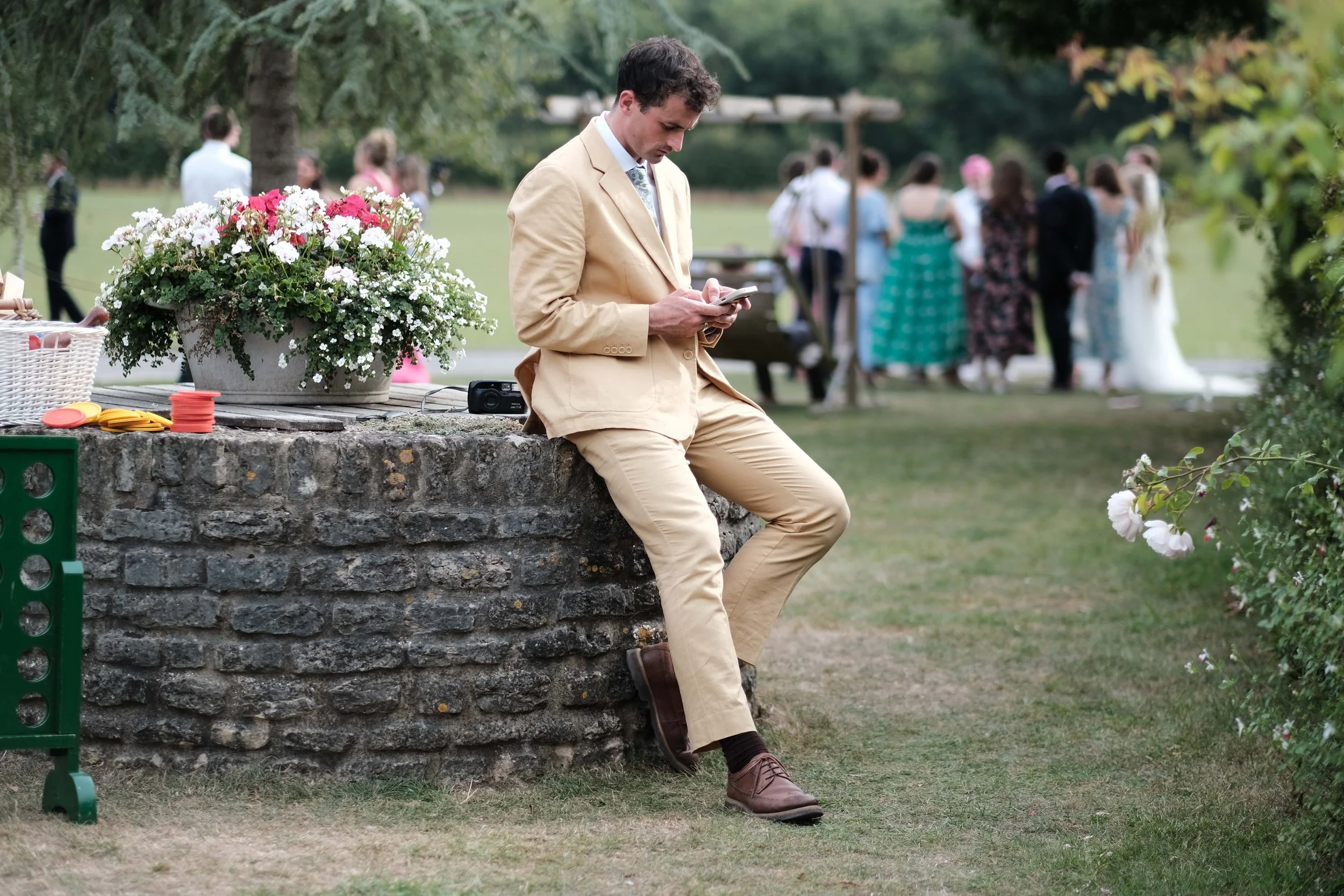 A man in a beige suit sitting on a stone ledge, looking at his phone, at an outdoor gathering or wedding event with people in the background.