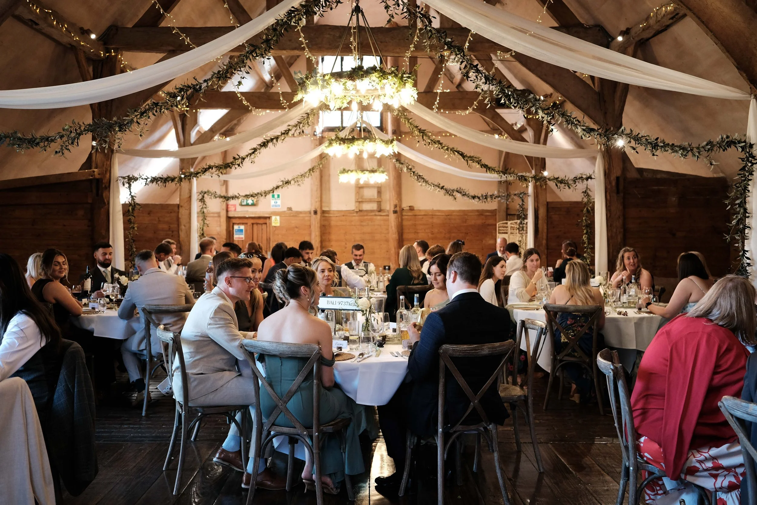 Wedding reception inside a rustic barn with wooden beams and hanging greenery and fairy lights, guests sitting around round tables with white tablecloths, engaged in conversations and enjoying a meal.