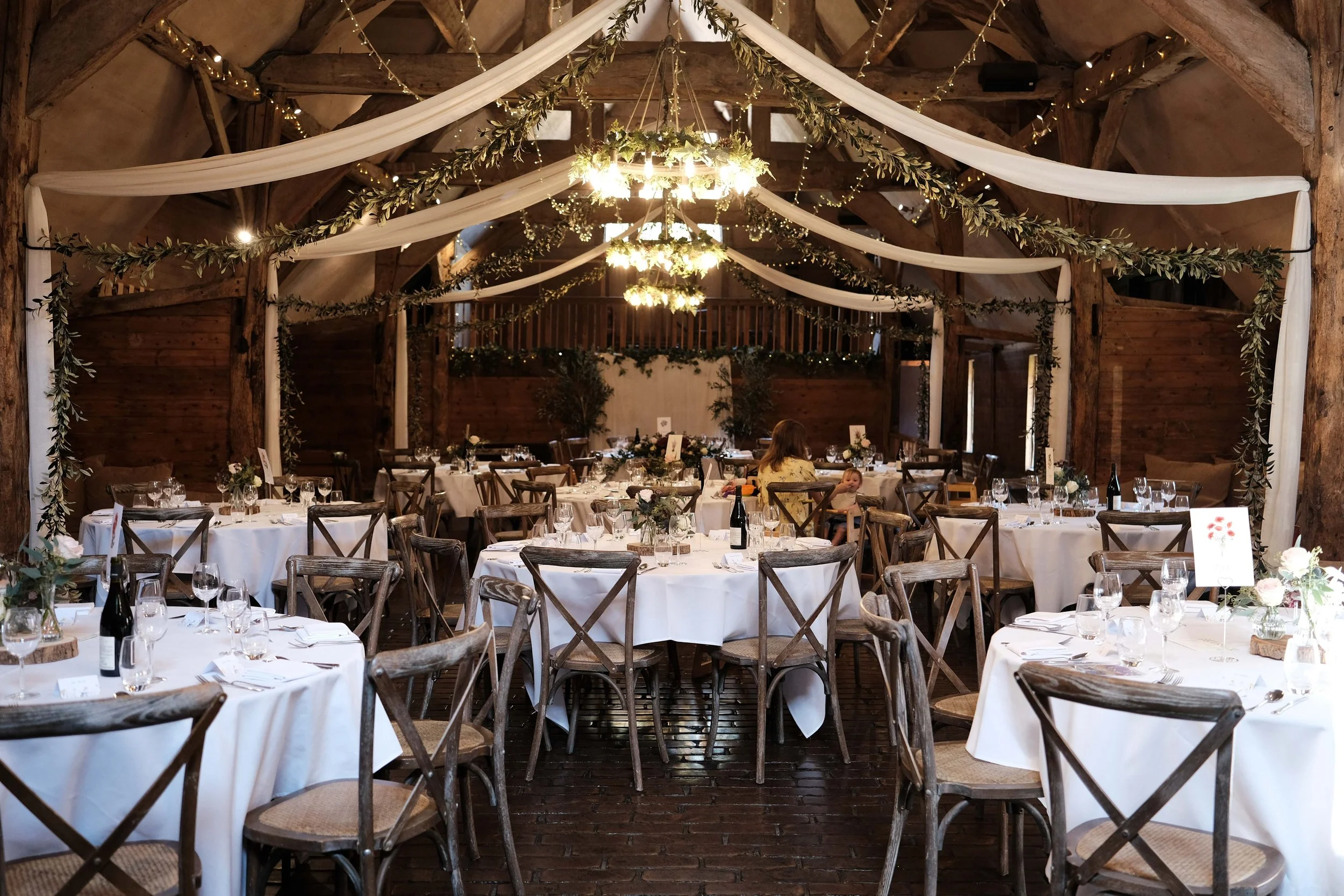 Decorated rustic wedding reception venue with round tables covered in white tablecloths, wooden chairs, floral centerpieces, wine bottles, and glassware, inside a barn with draped fabric and string lights.