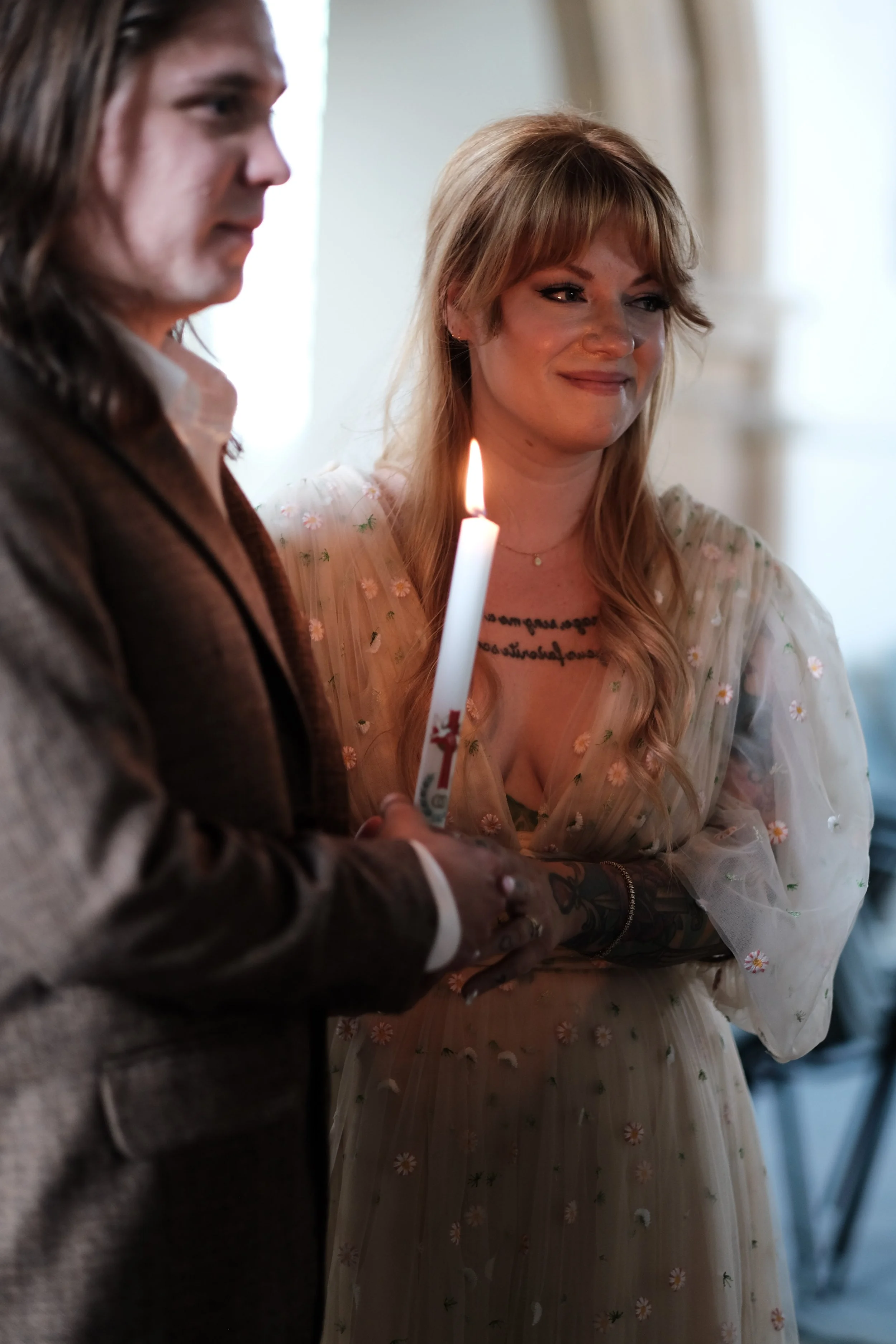A woman with long reddish hair and tattoos, wearing a sheer floral dress, holding a lit white candle, standing next to a man with long hair in a suit. They are indoors with a blurred background.