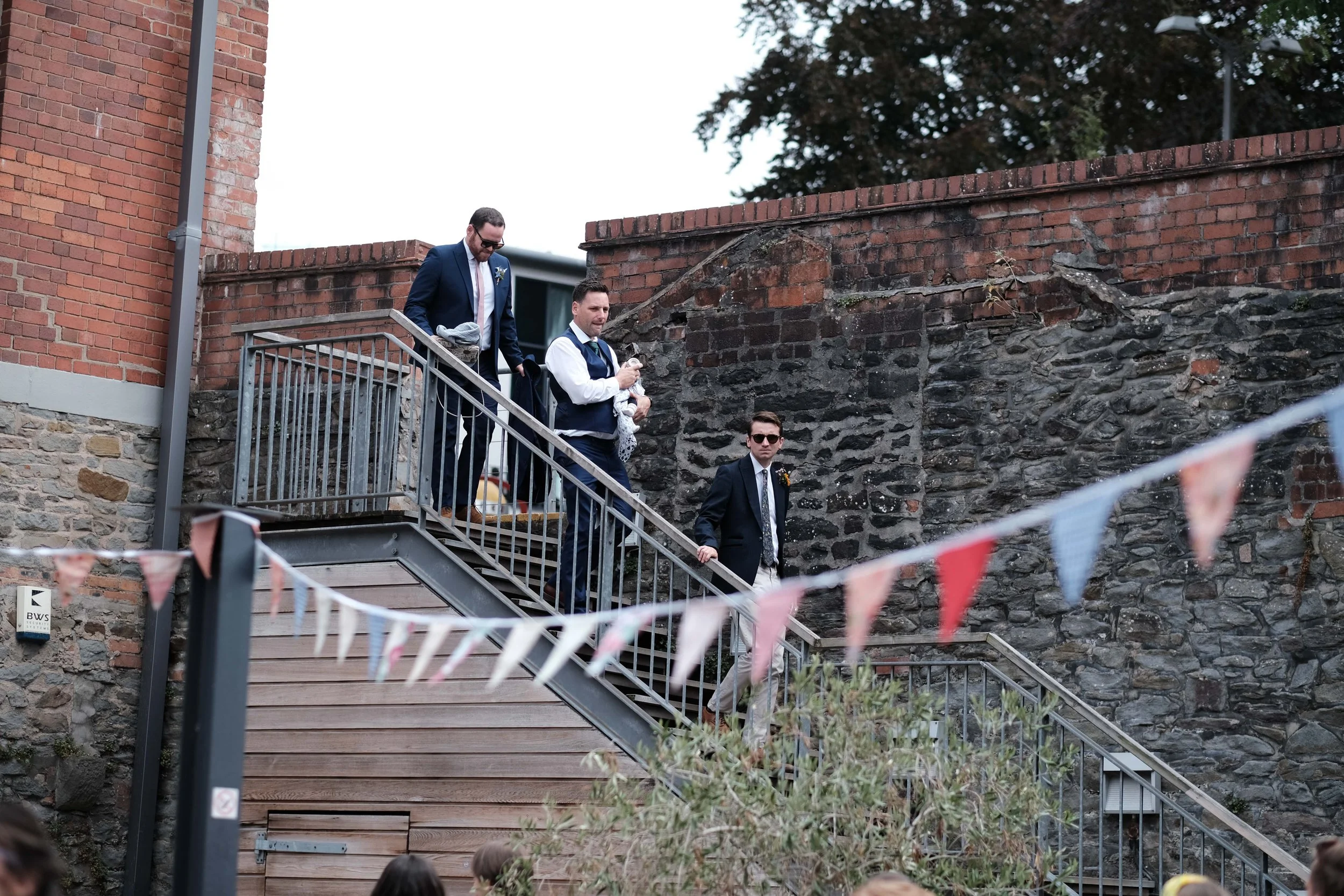 Three men descending outdoor staircase, one holding a baby, near stone and brick wall, with pink and blue bunting.