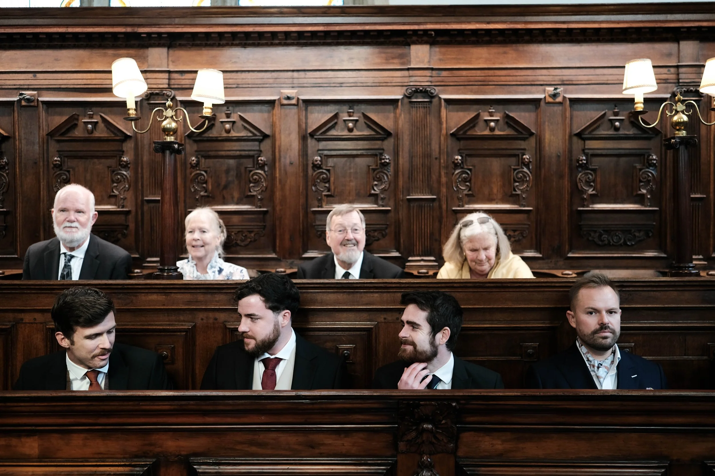Seven people sitting in a wooden courtroom or legislative chamber. Four older people at the top and three younger men at the bottom, all dressed in formal attire.