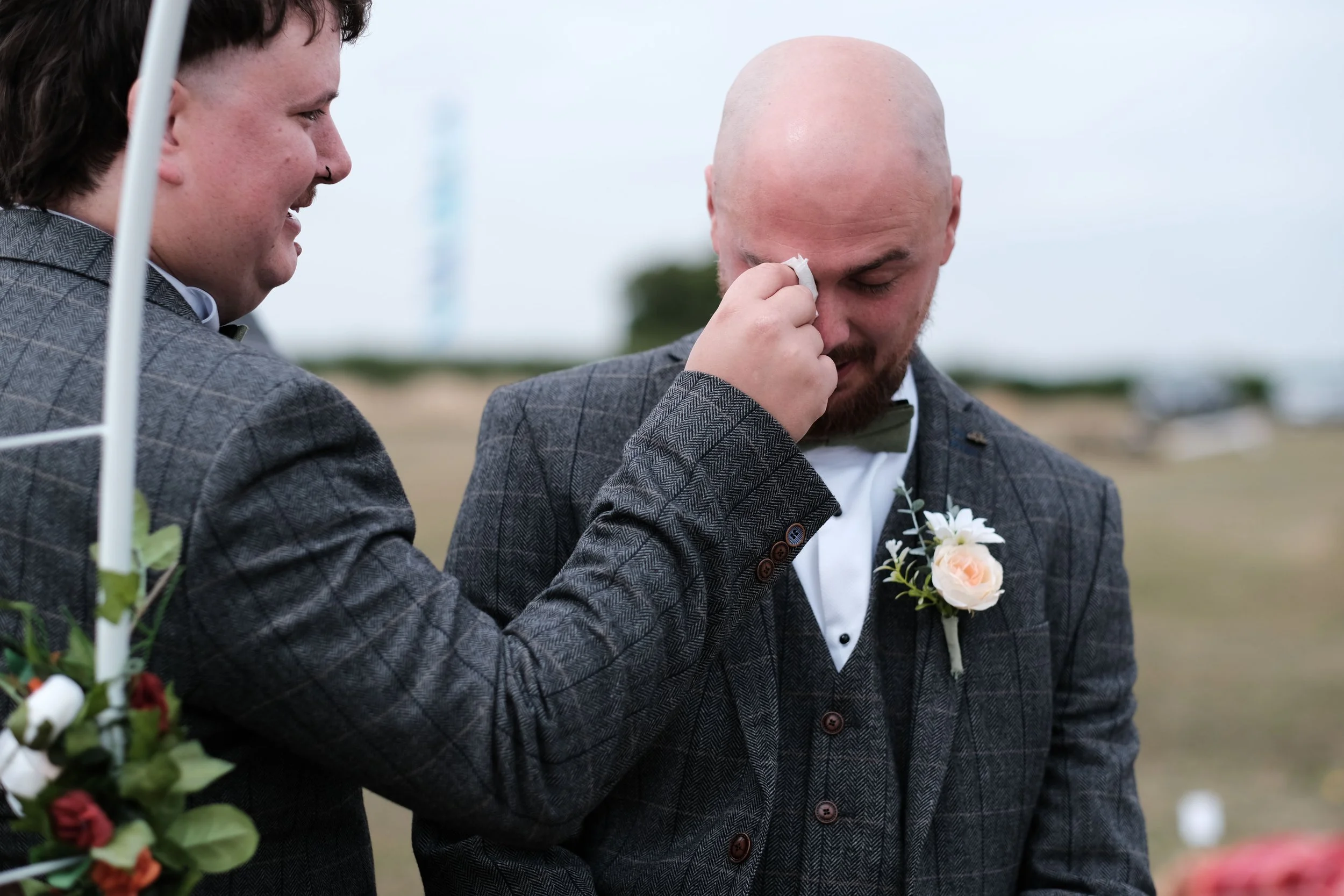 A man with a beard is wiping tears from his eyes during a wedding ceremony, wearing a gray checkered suit with a white boutonnière. Another man in a similar suit is smiling beside him, holding a bouquet of flowers.