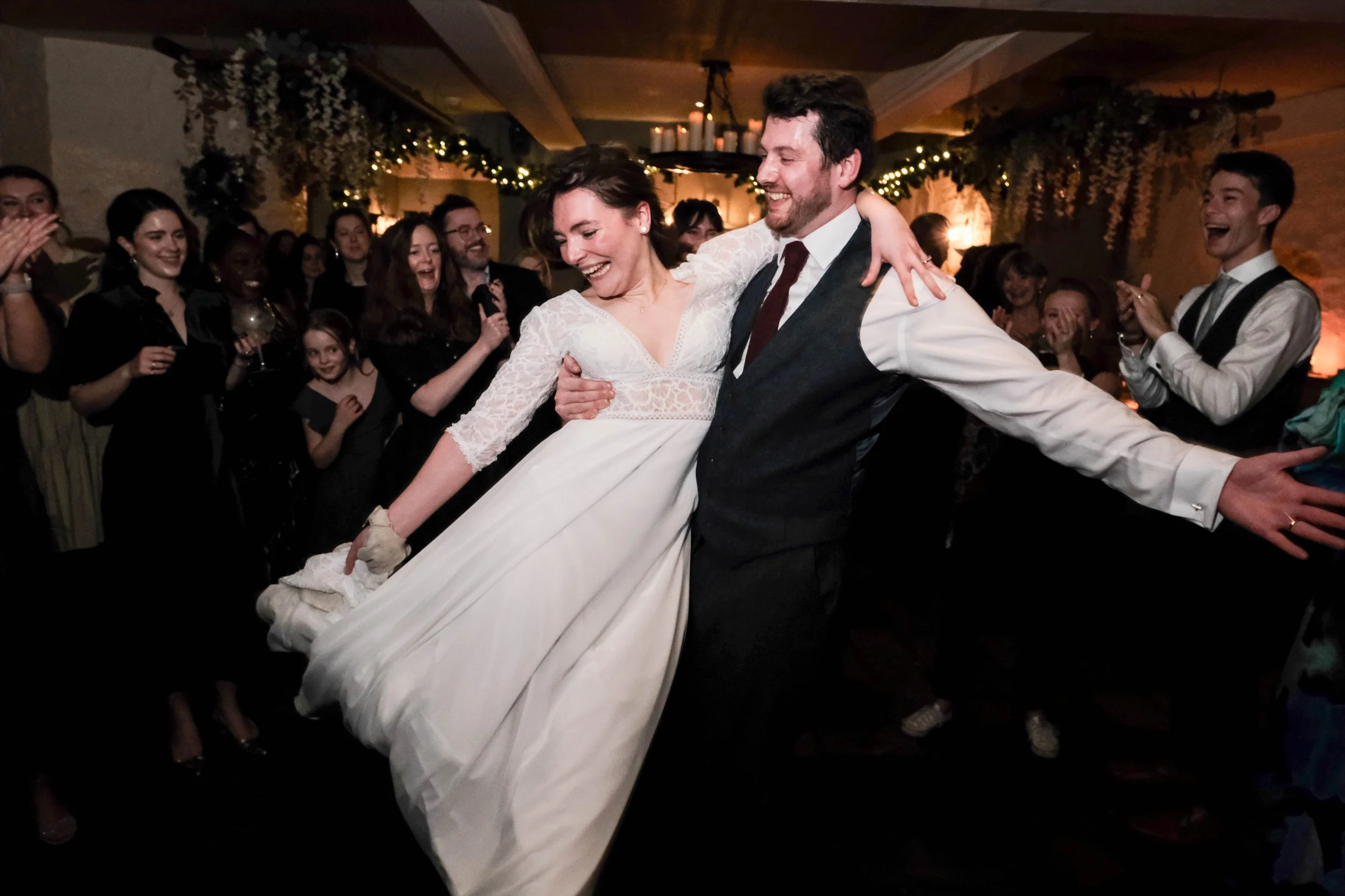 Couple dancing at wedding reception, surrounded by guests clapping and smiling, with festive decorations and warm lighting.