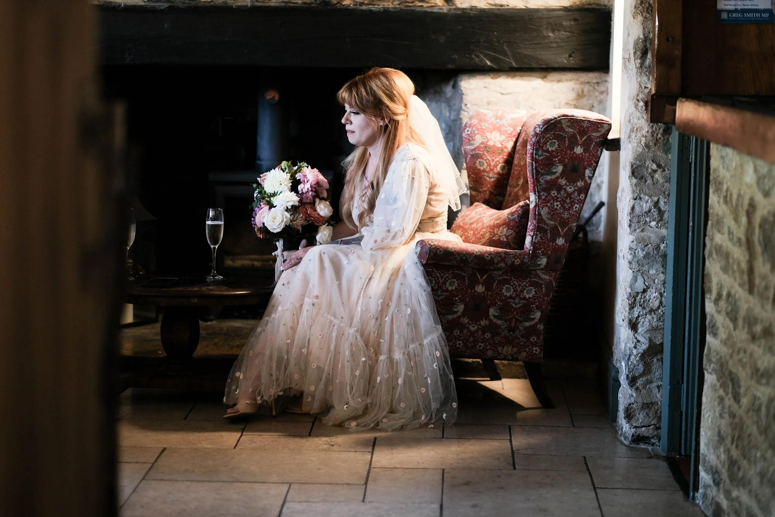 A woman with long red hair in a white dress sitting on a floral armchair in a rustic room, holding a bouquet of flowers, with a glass of champagne on a small table beside her.