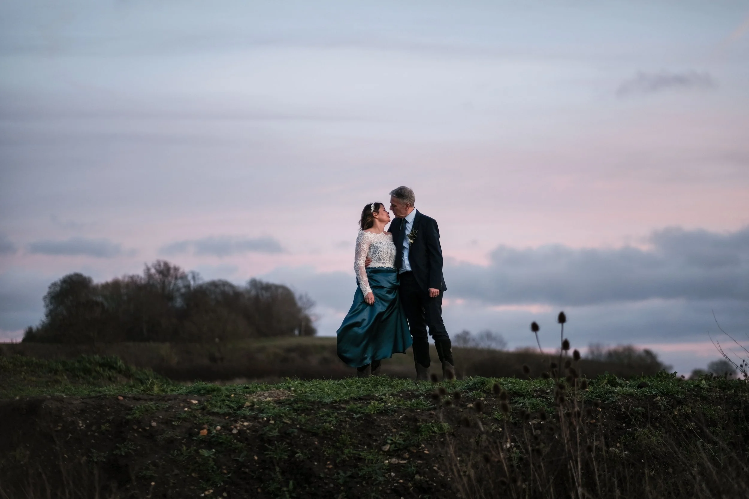 A couple dressed in formal attire, a woman in a white lace top and teal skirt, and a man in a black suit, sharing a romantic moment outdoors at sunset with a cloudy sky and trees in the background.