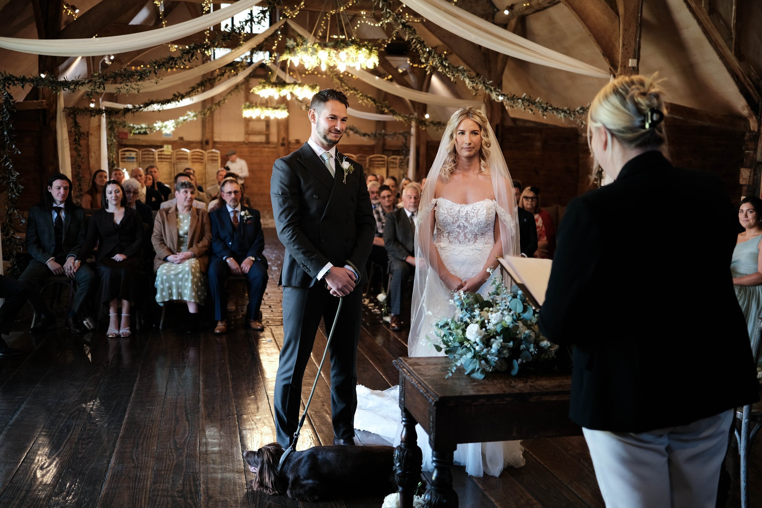A wedding ceremony taking place inside a rustic barn with indoor lighting and draped white fabric and green foliage decorations. The bride and groom stand before an officiant, with the groom holding a leash attached to a small dog. The bride is in a 
