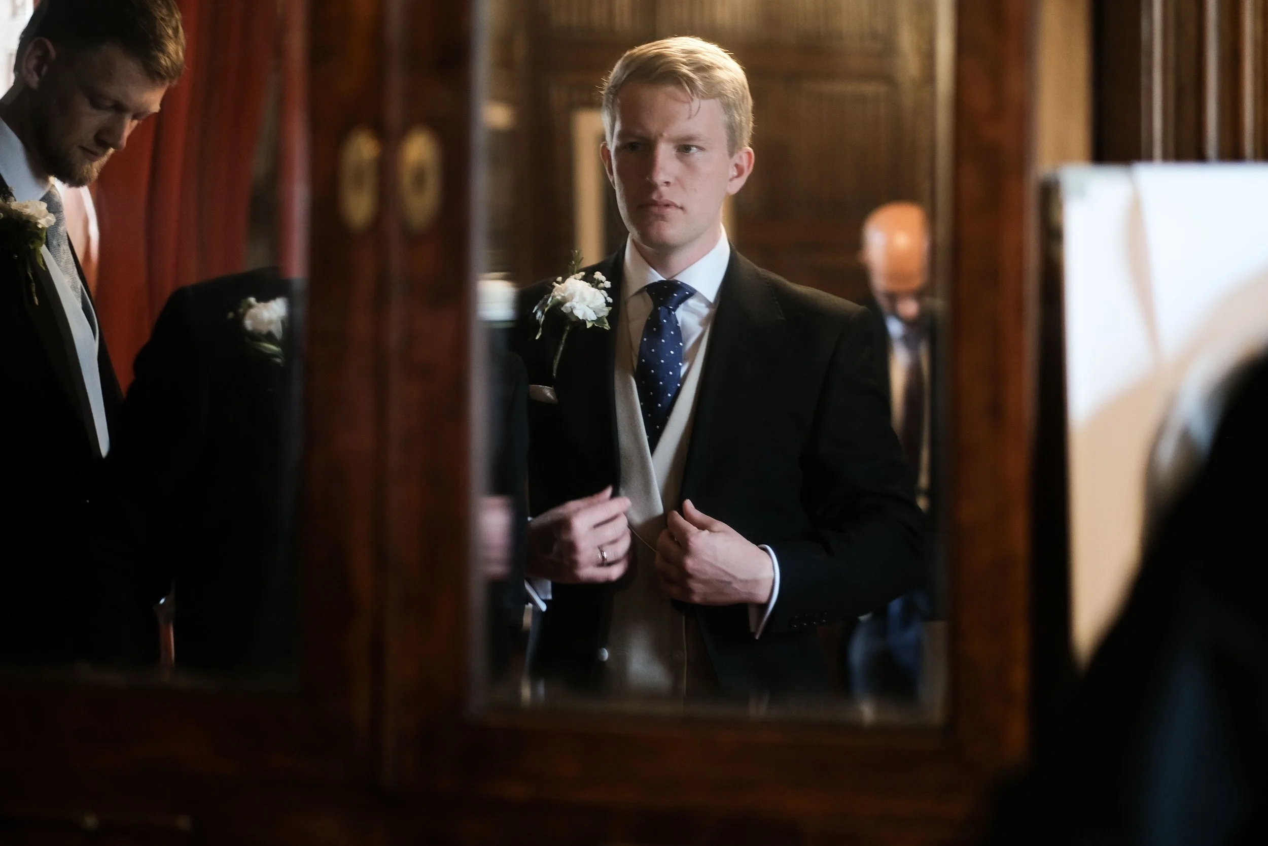 A young man in a black suit, white shirt, and blue polka dot tie, adjusting his jacket, seen through an ornate wooden mirror at a formal event or wedding. Other people and wood-paneled walls are visible in the background.