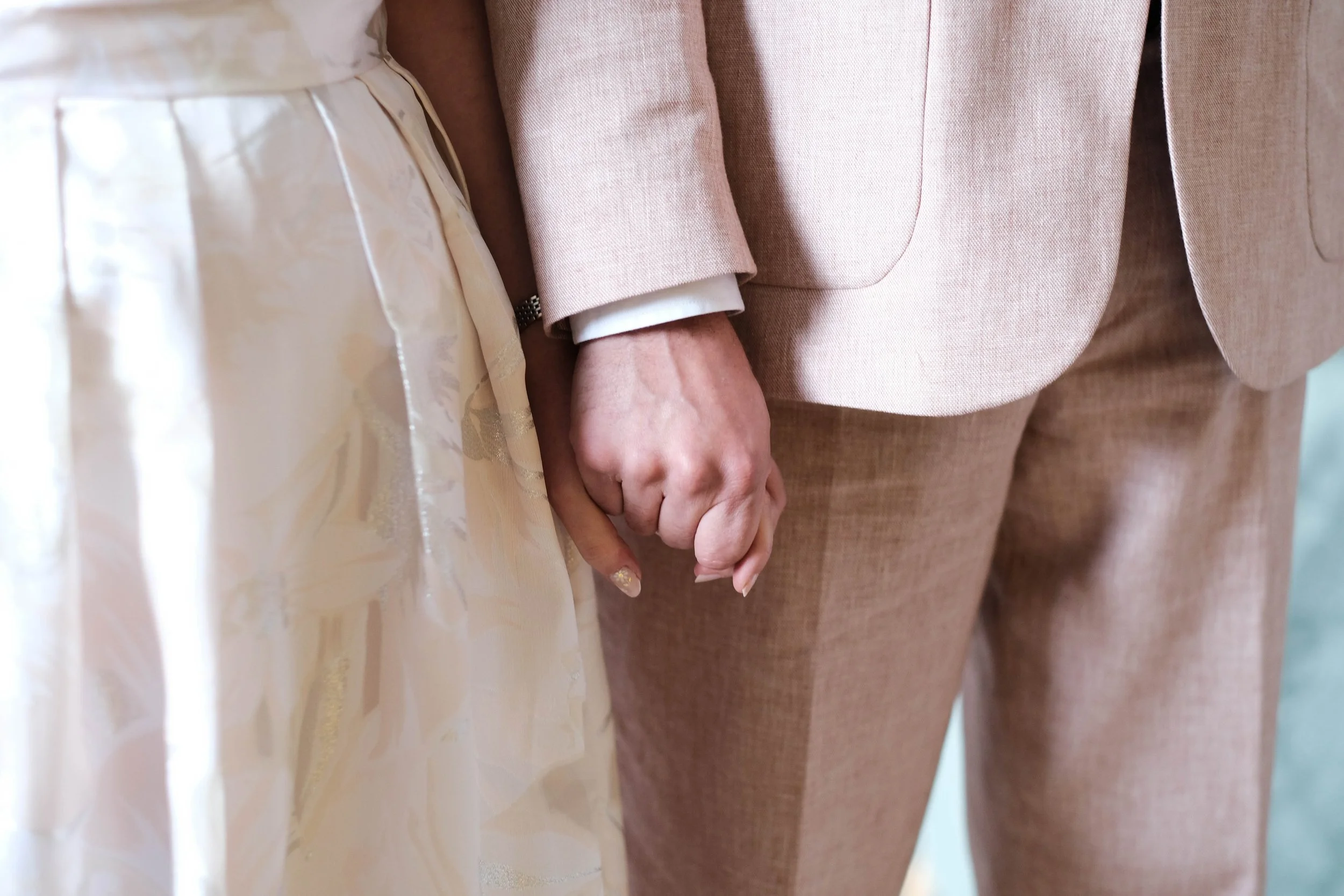 Close-up of a couple holding hands, with the woman wearing a white wedding dress and the man in a beige suit.