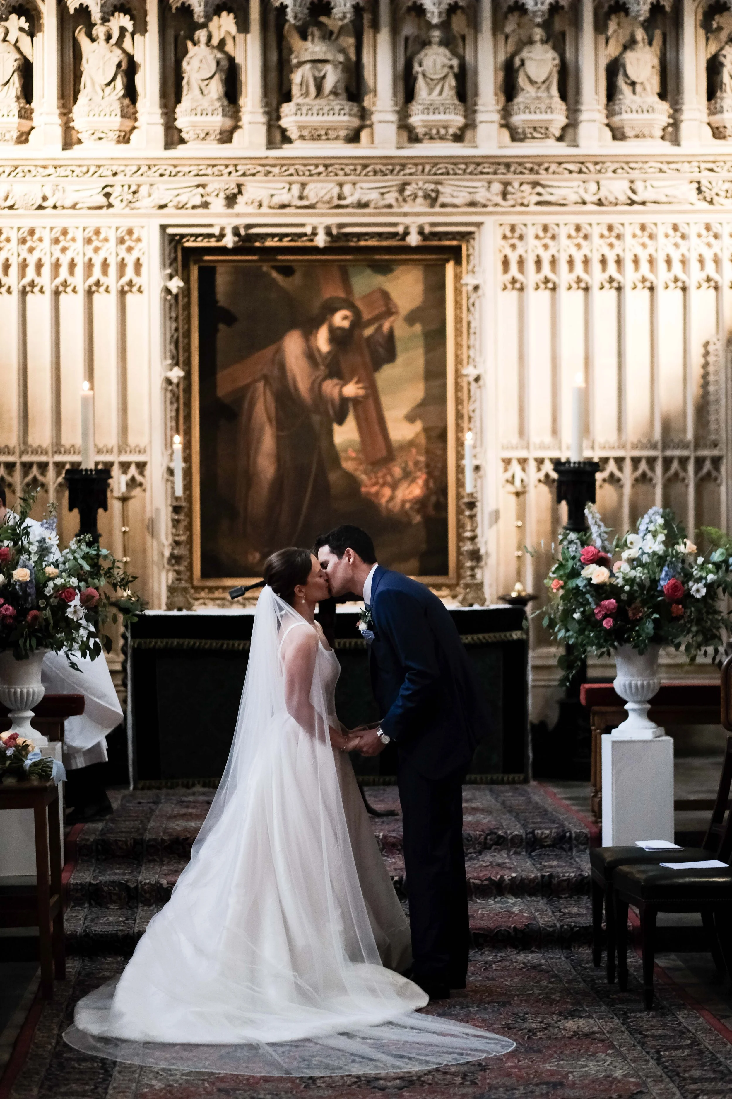 A bride and groom kissing during their wedding ceremony inside a church with an ornate altar and religious paintings in the background.