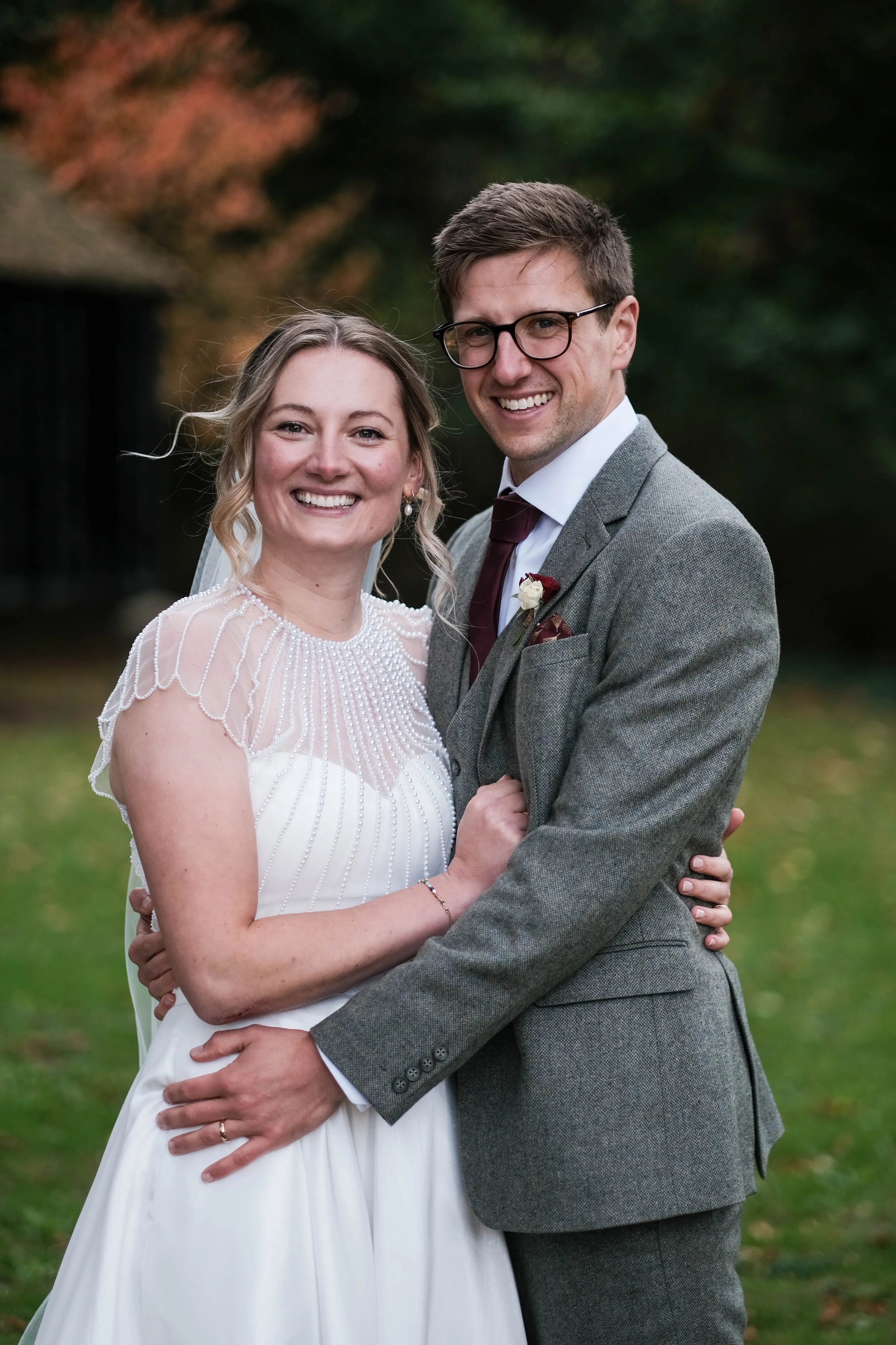 A smiling bride and groom embrace outdoors. The bride wears a white wedding dress with beaded detailing on the shoulders. The groom wears a grey suit, white shirt, and maroon tie. They stand on a grassy area with blurred trees in the background.