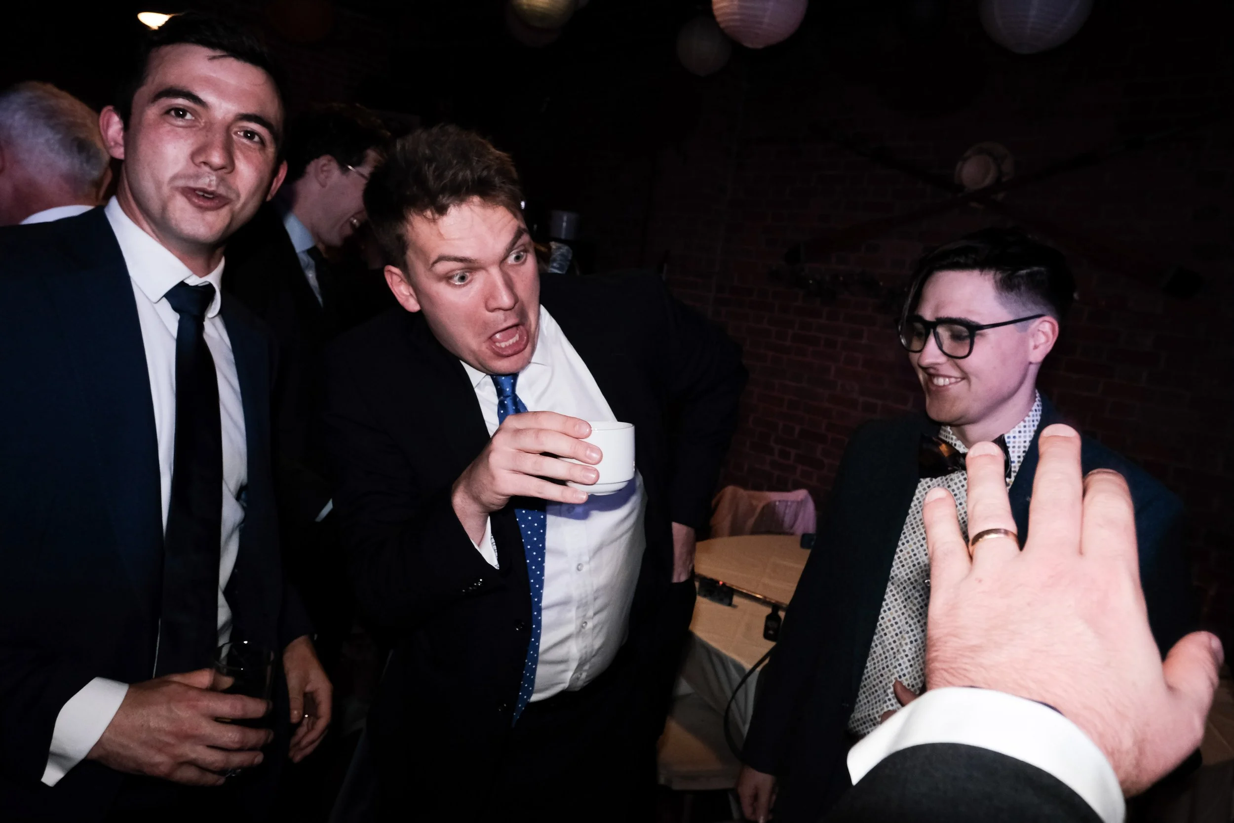 Group of men in formal attire socializing at a party, with brick wall background.