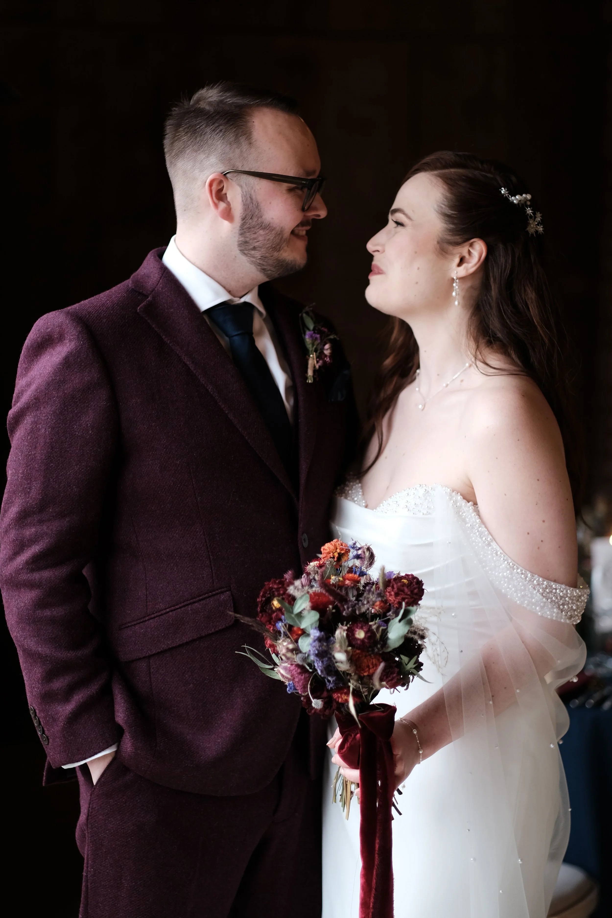 A bride and groom stand close, facing each other and smiling. The bride is in a white gown holding a bouquet, and the groom is in a dark suit with a boutonniere, both smiling.