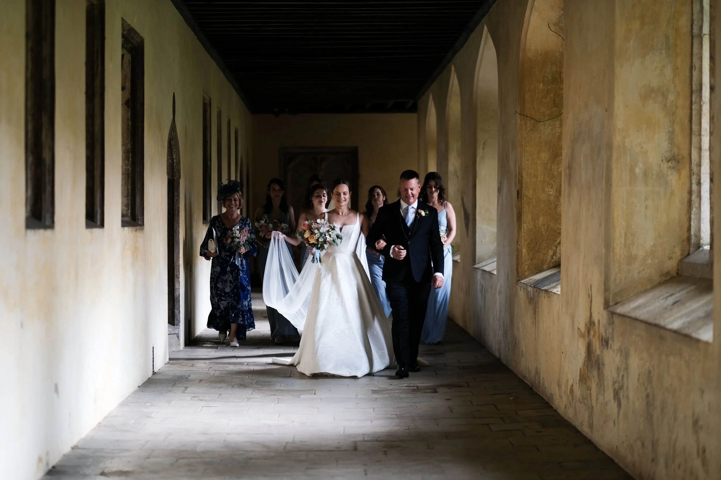 Wedding procession of bride and groom walking through a historic corridor, surrounded by bridesmaids and family.