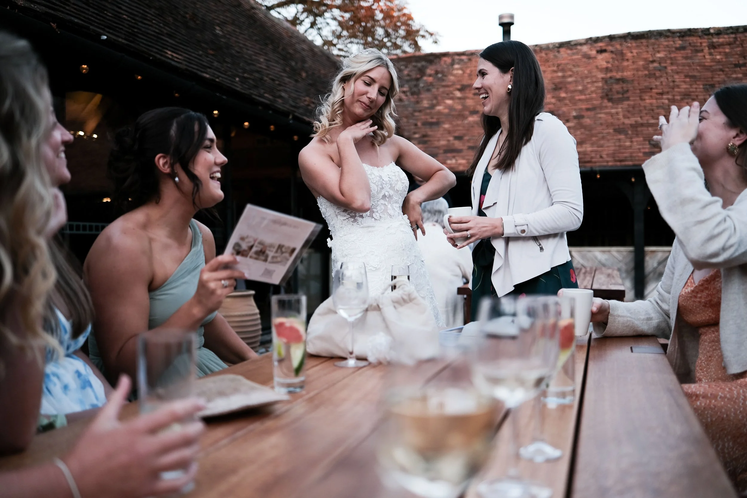 Group of women celebrating at a wedding reception outdoors, with the bride in a white gown standing and smiling as she listens to a woman in a white blazer holding a cup. Other women are seated around the table holding drinks and smiling.