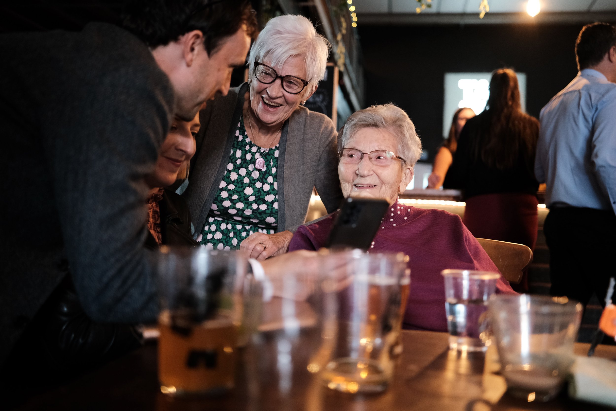 Group of people, including an elderly woman with glasses and a woman with short white hair, looking at a smartphone in a social setting