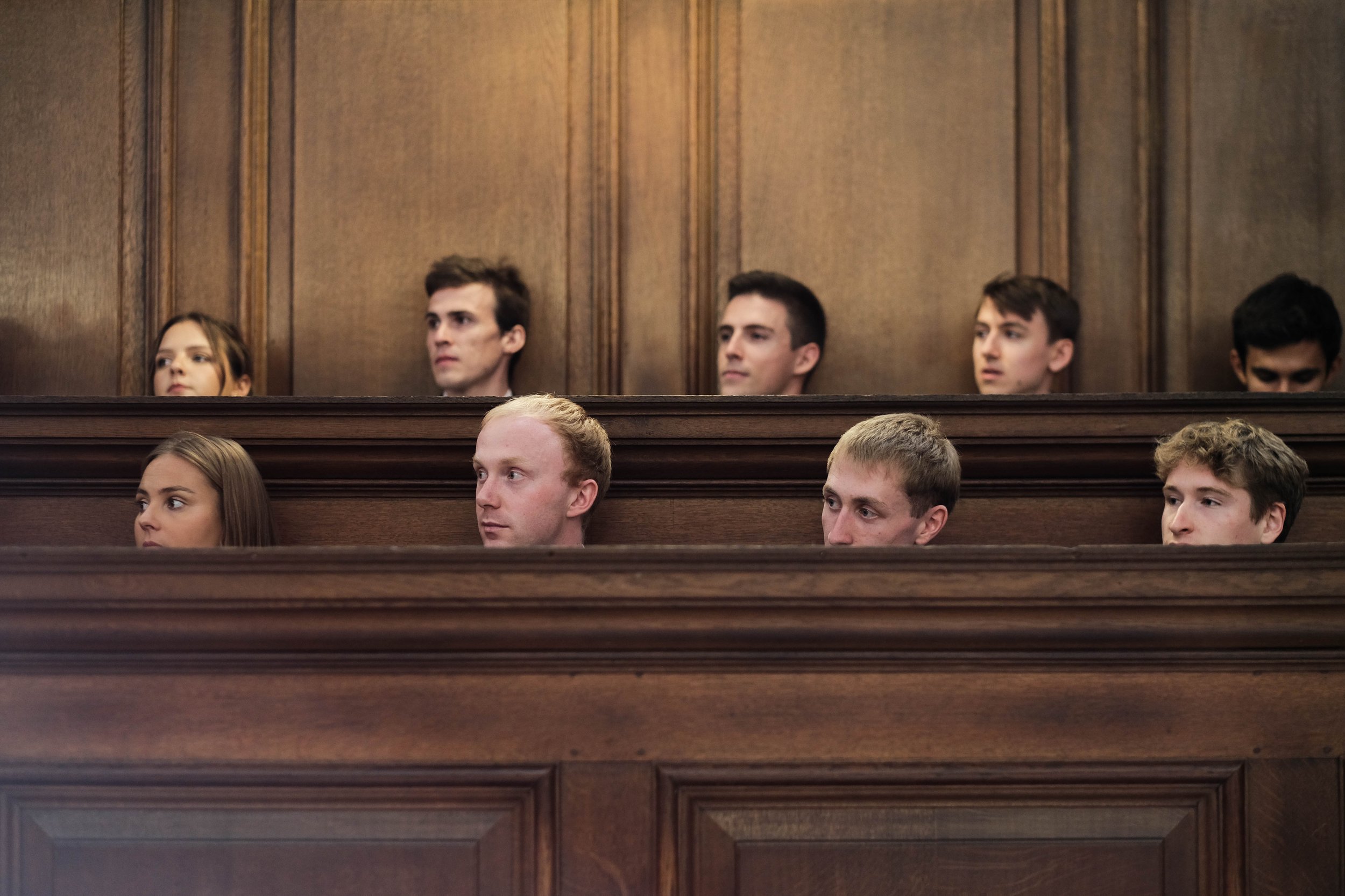 People sitting in a courtroom with wooden paneling, with some visible faces in the gallery.