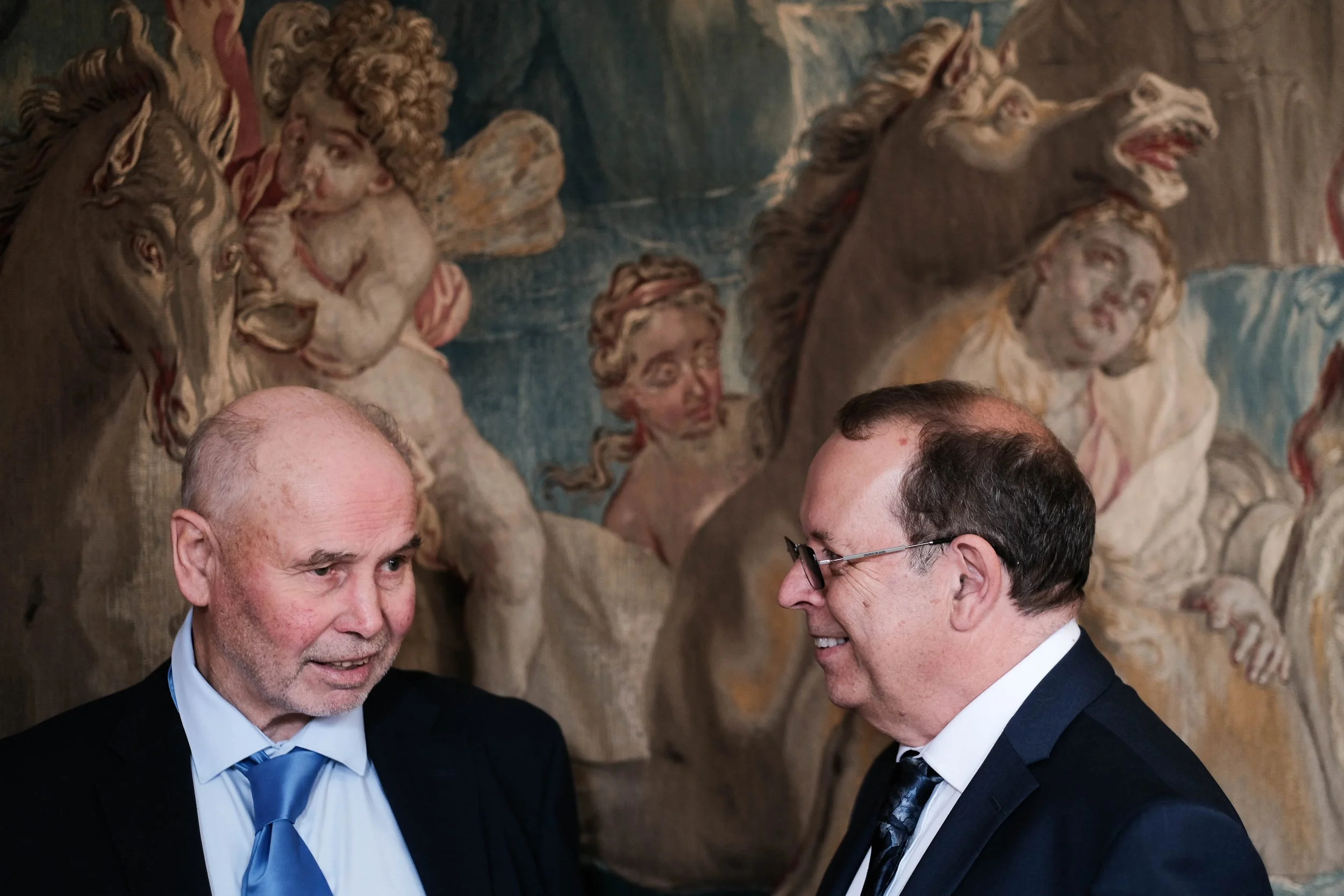 Two men in suits talking in front of a mural with mythological figures, including winged children and horse-headed creatures.
