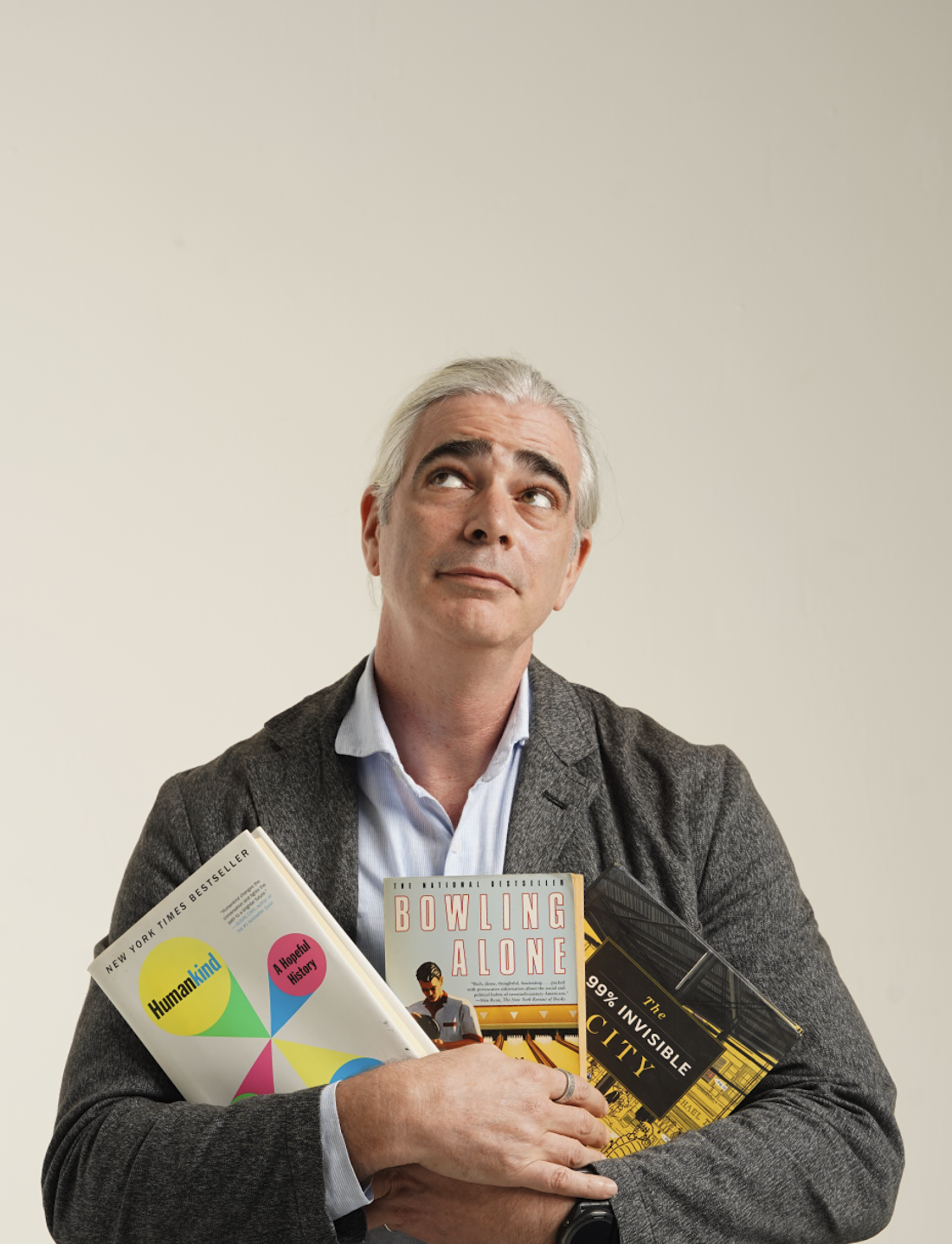 A middle-aged man with gray hair, wearing a gray blazer and blue shirt, holding three books titled 'Humankind,' 'Bowing Alone,' and 'The 99% Invisible City,' standing against a plain light-colored background, looking up and slightly to the side.