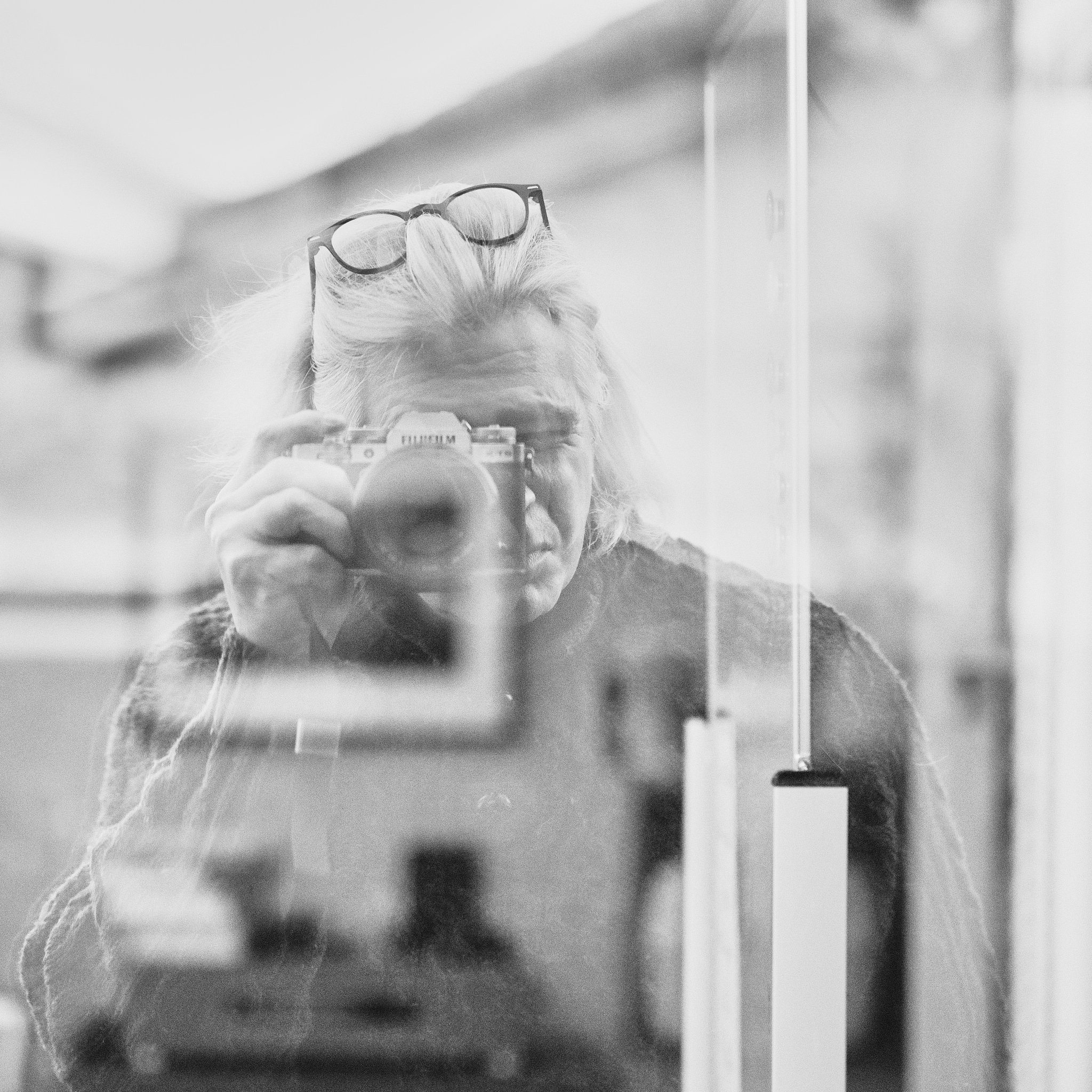 A person taking a mirror selfie over their shoulder with glasses resting on their head, visible hand holding a camera, and blurred background.