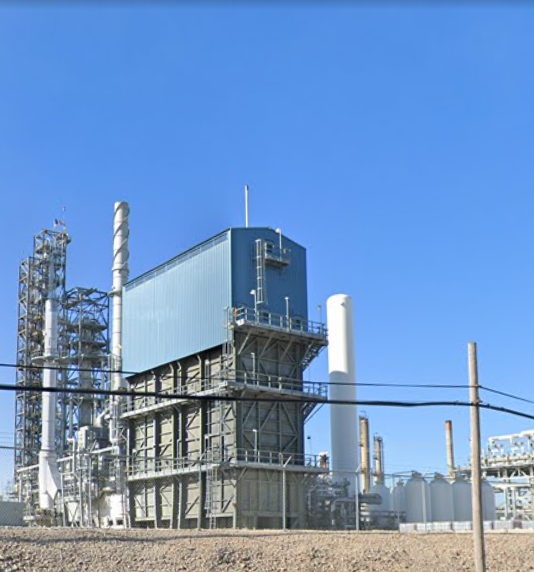 Industrial factory or power plant with metal structures, pipes, and a blue building under a clear blue sky.