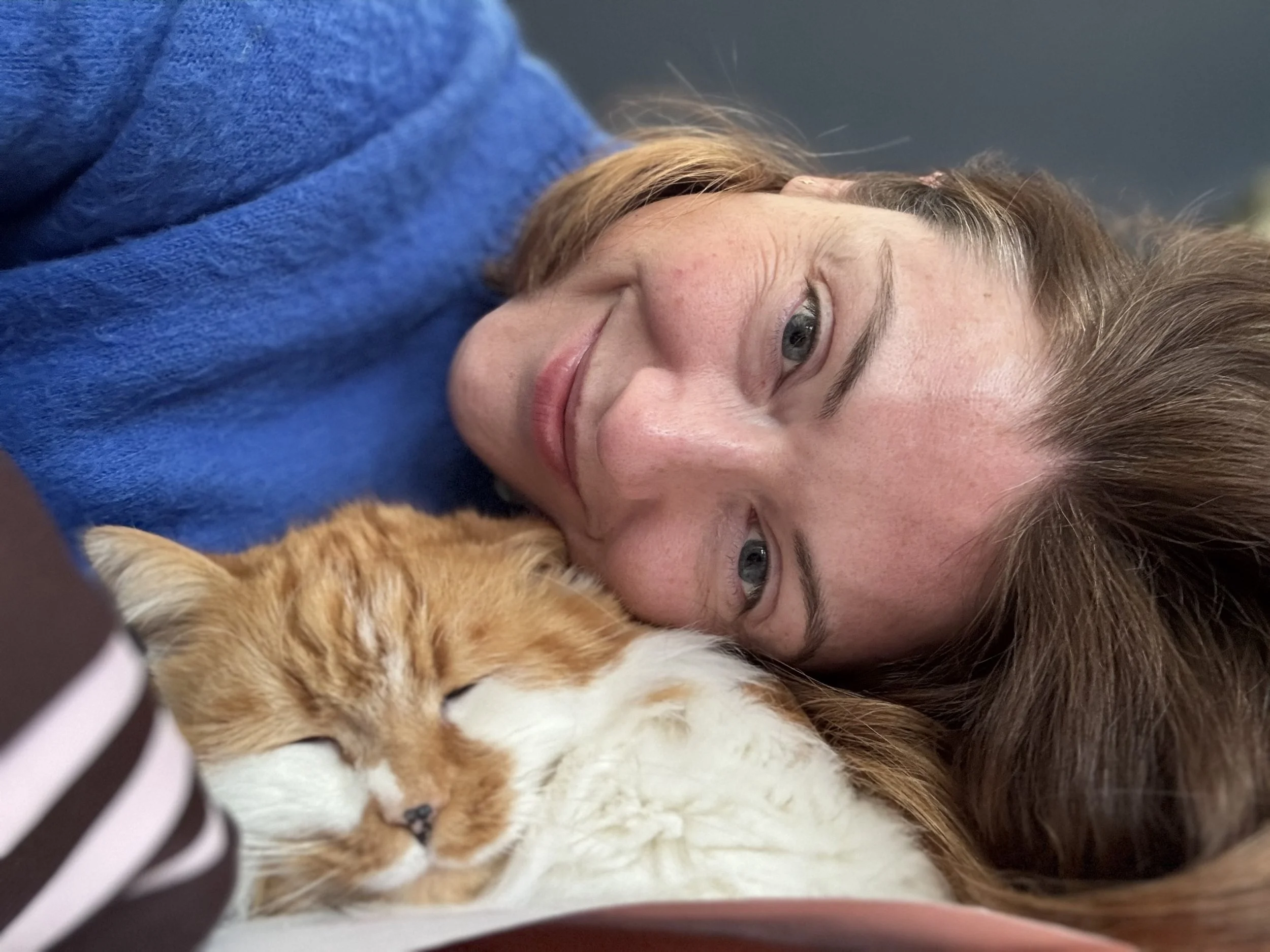 A woman with red hair lying next to a sleeping orange and white cat, both resting on a pillow.