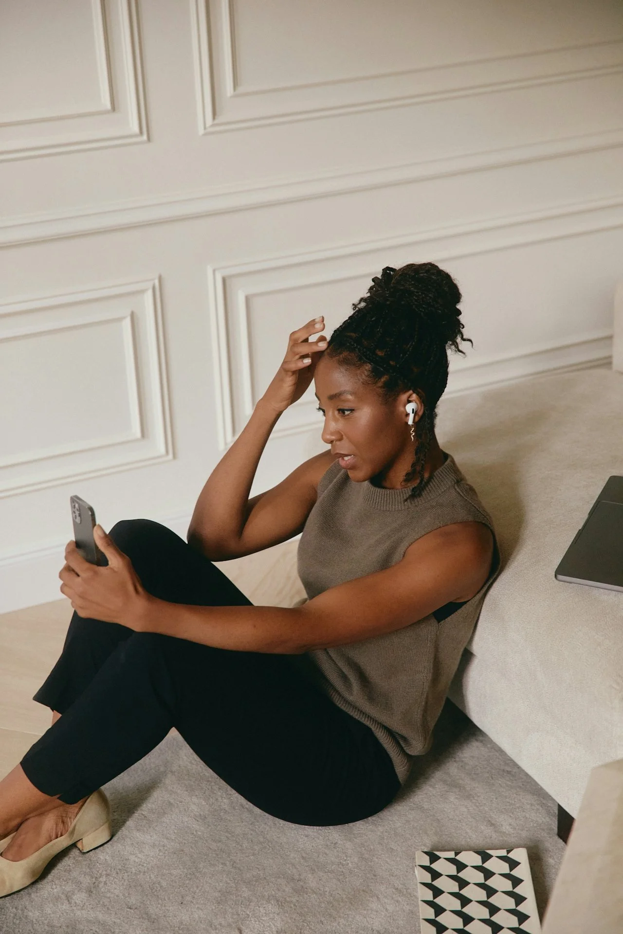 A woman with braided hair tied up in a bun, sitting on the floor next to a beige sofa, looking at her smartphone, wearing wireless earbuds, a sleeveless sweater, black pants, and beige heeled shoes.