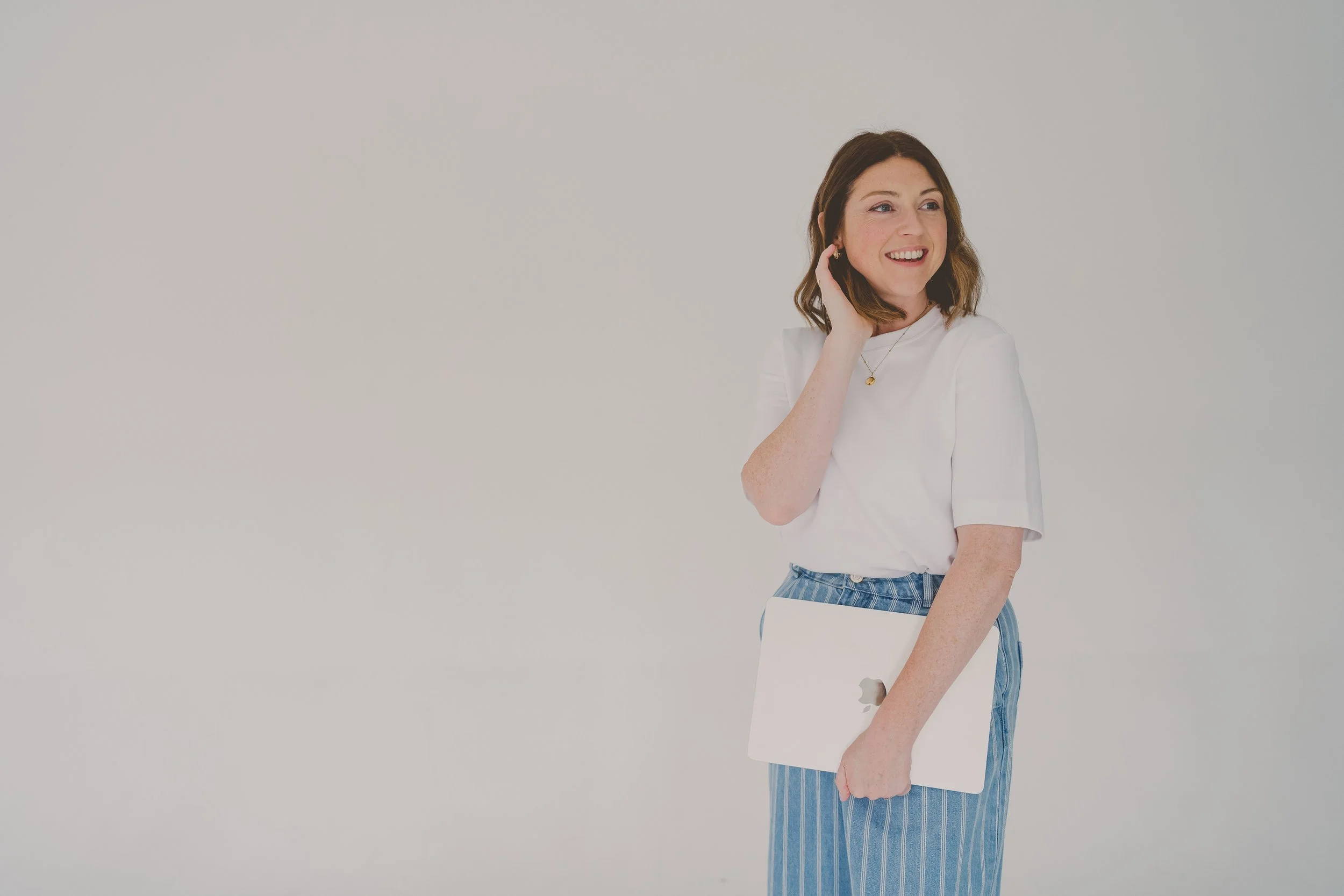 A woman with shoulder-length wavy brown hair, wearing a white T-shirt and blue striped pants, holding a closed silver laptop with an Apple logo, smiling and touching her hair against a plain white background.