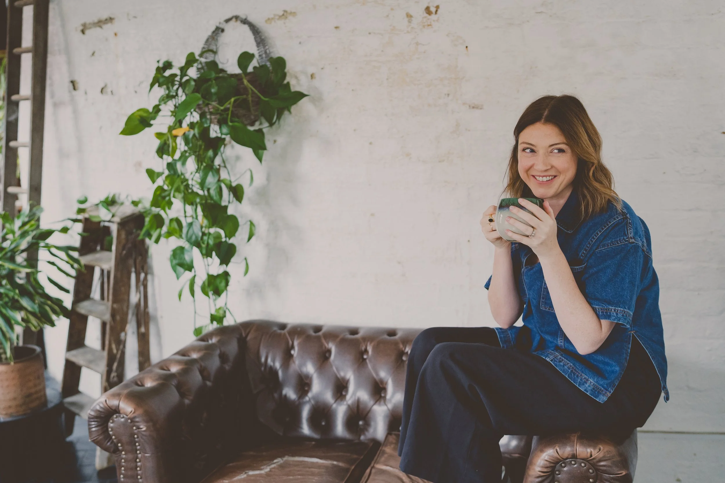 A woman sitting on a brown leather tufted sofa holding a mug, smiling and looking at the camera.