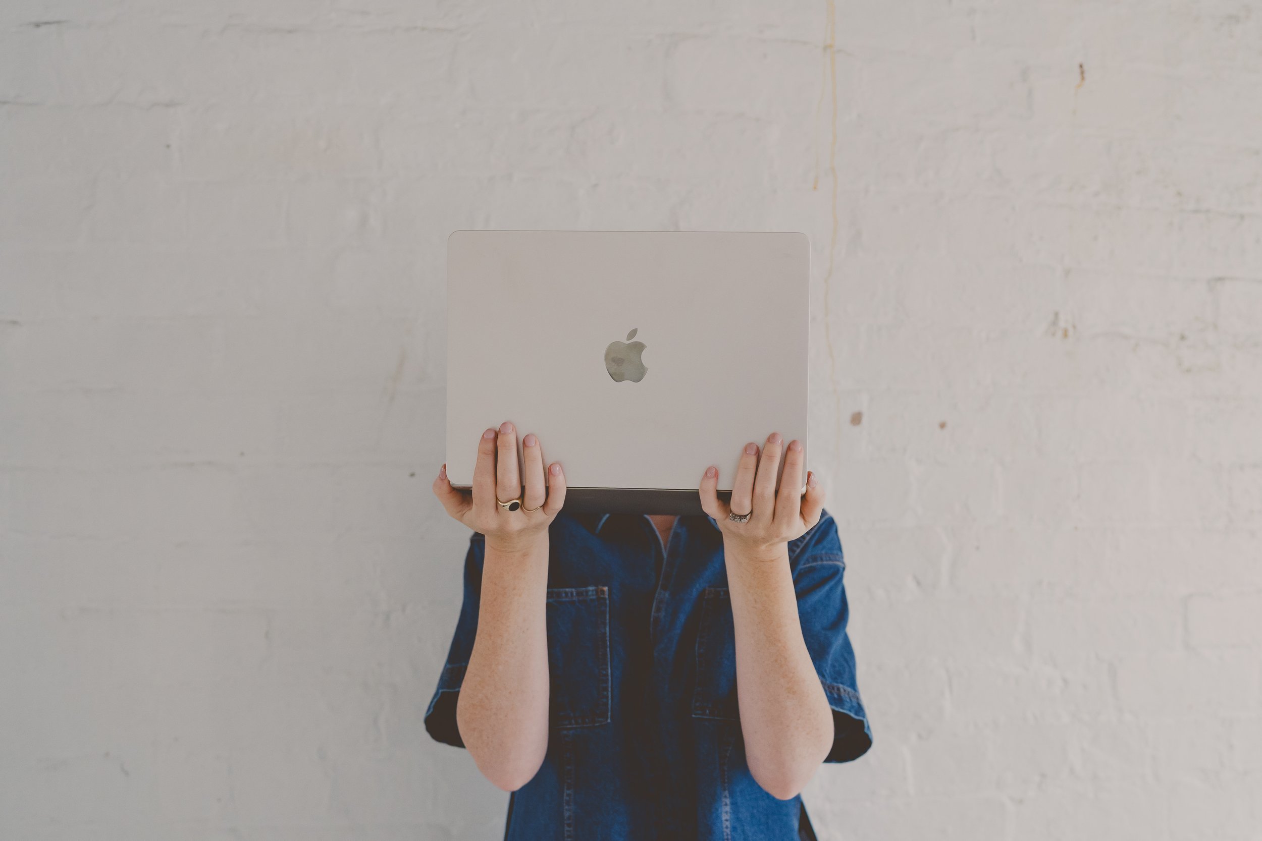 Person holding a silver MacBook in front of their face, wearing a denim shirt, against a white brick wall.