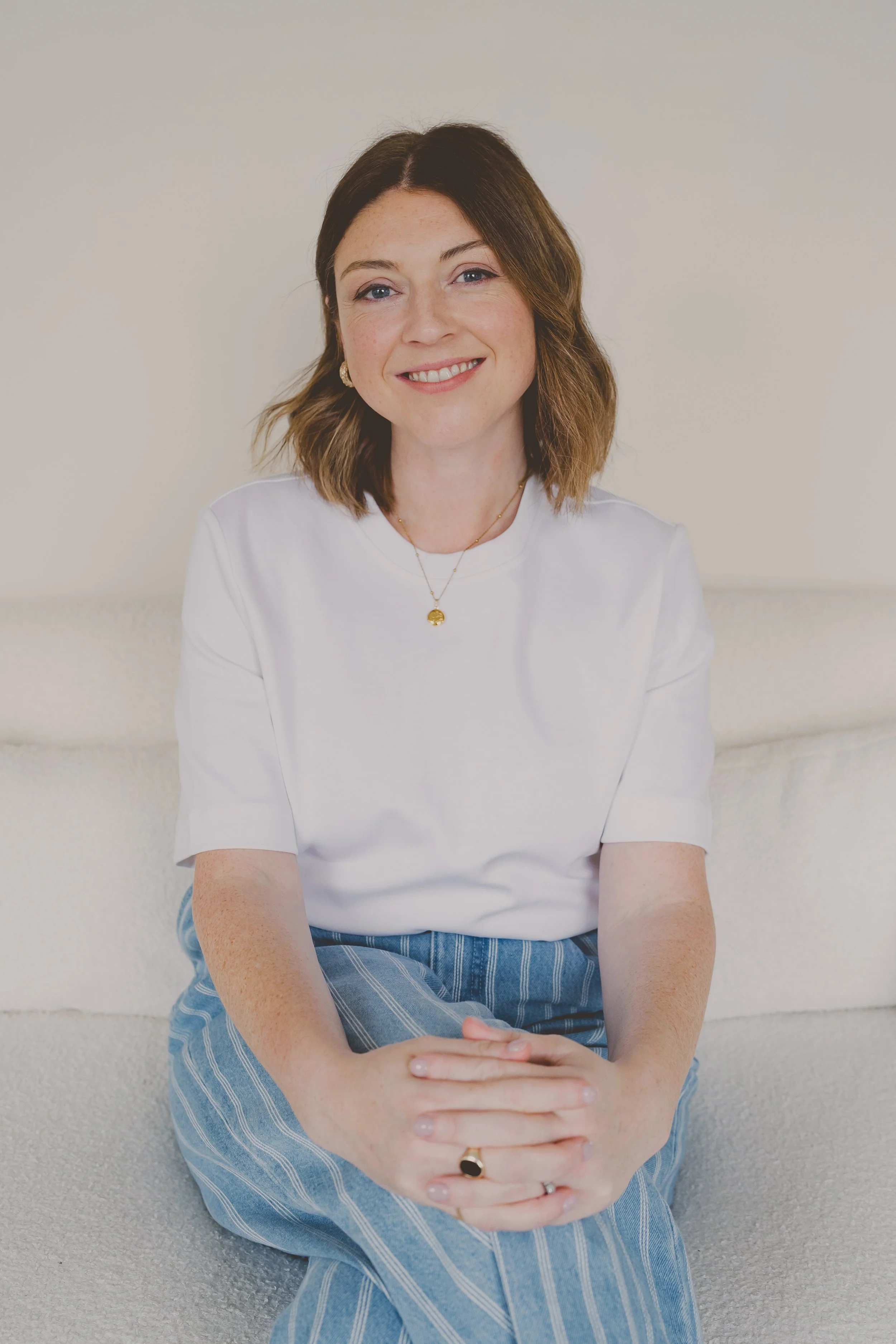 A woman with shoulder-length brown hair, wearing a white t-shirt and blue striped pants, is sitting on a white sofa, smiling at the camera.