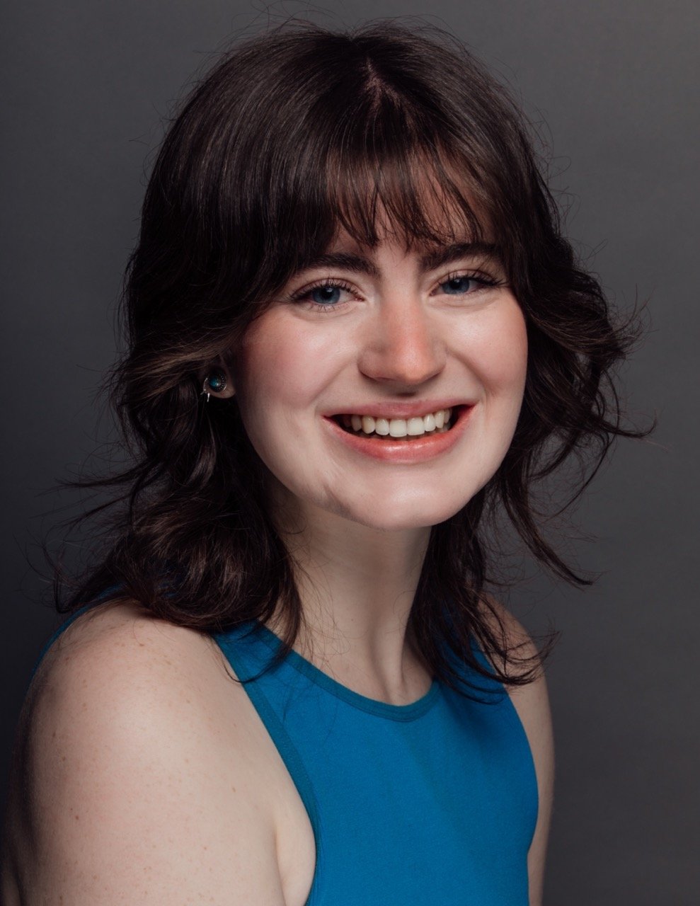 A young woman with curly brown hair, blue eyes, and light skin, smiling and wearing a blue sleeveless top, posed against a dark gray background.