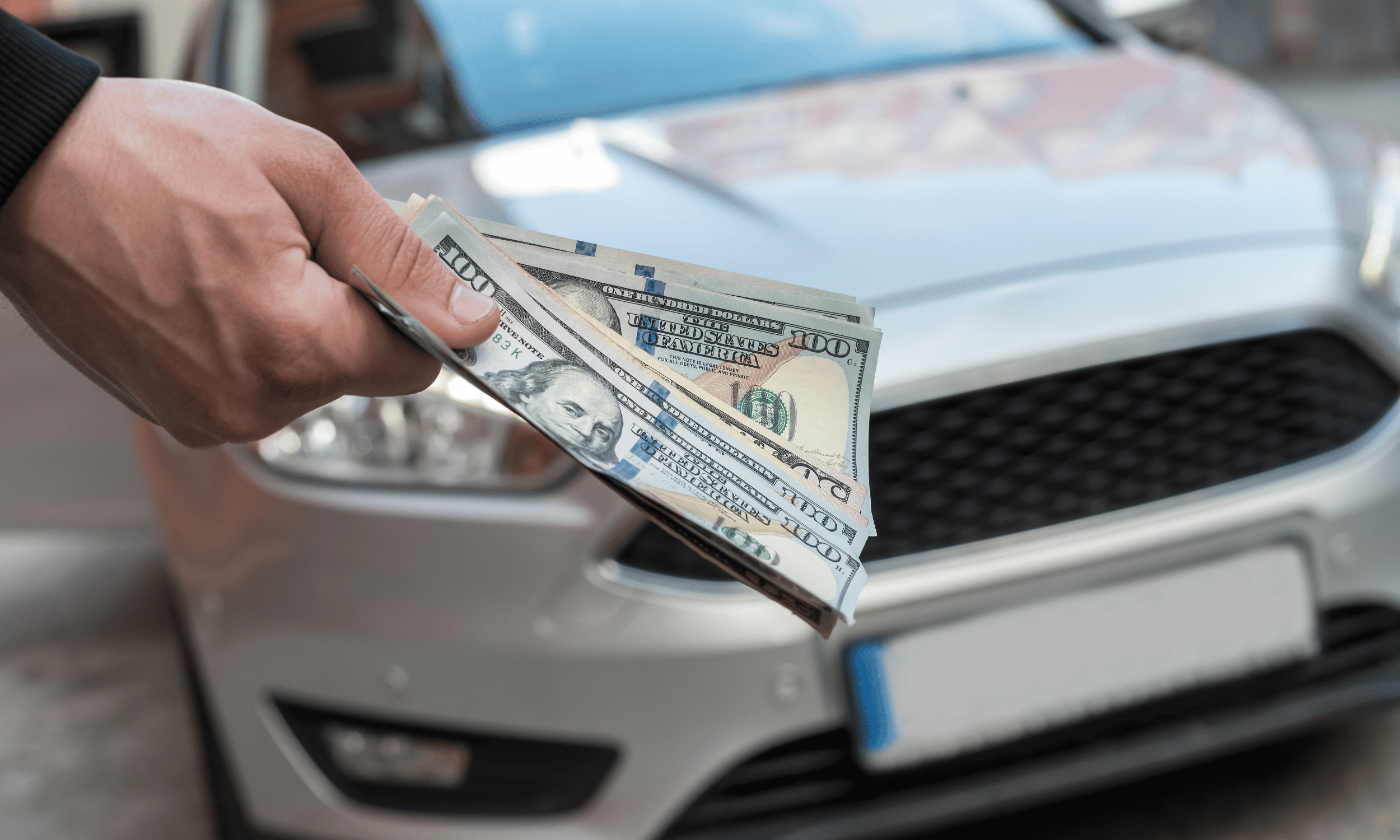 Hand holding US dollar bills in front of a car.