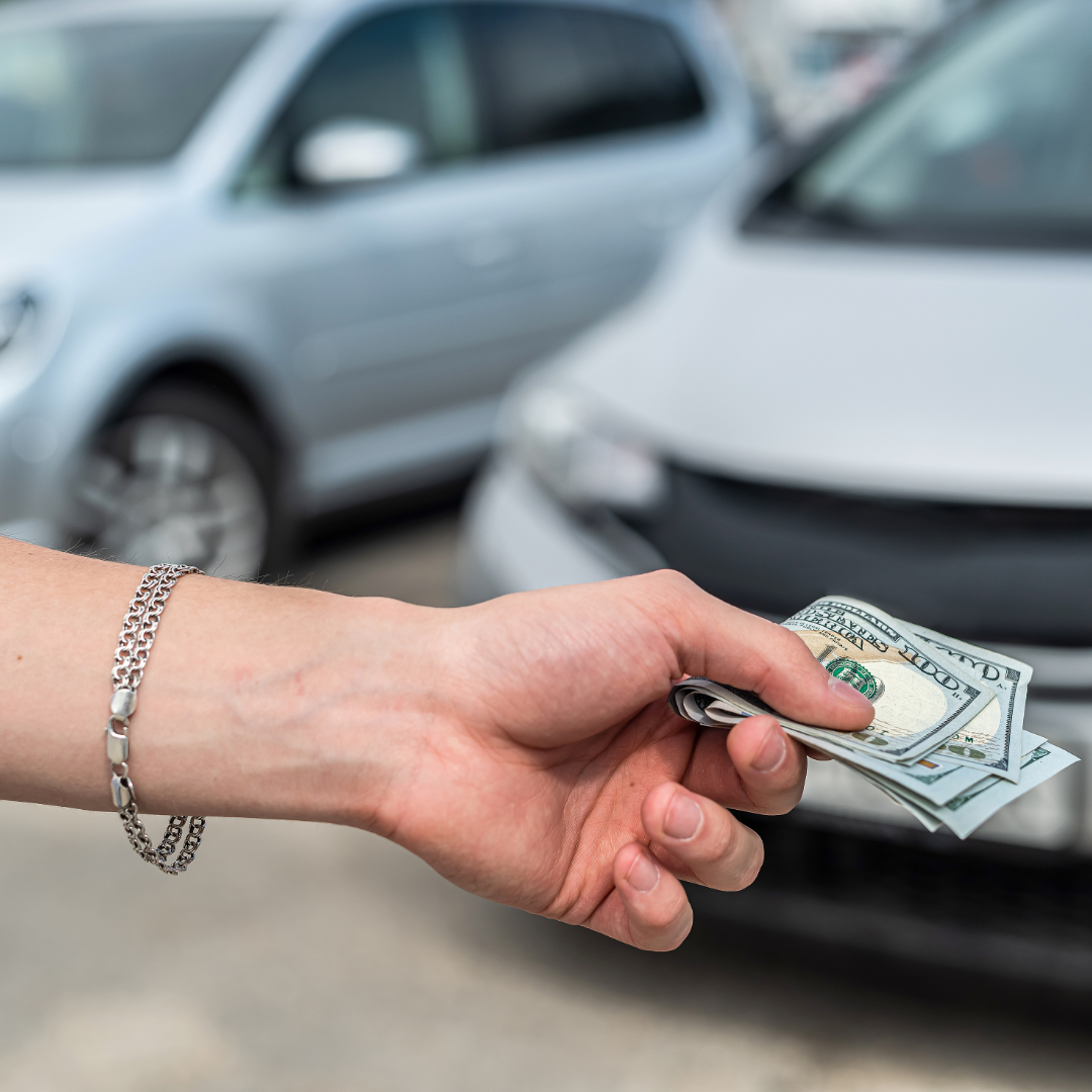 A person's hand holding a stack of hundred-dollar bills in front of parked cars.