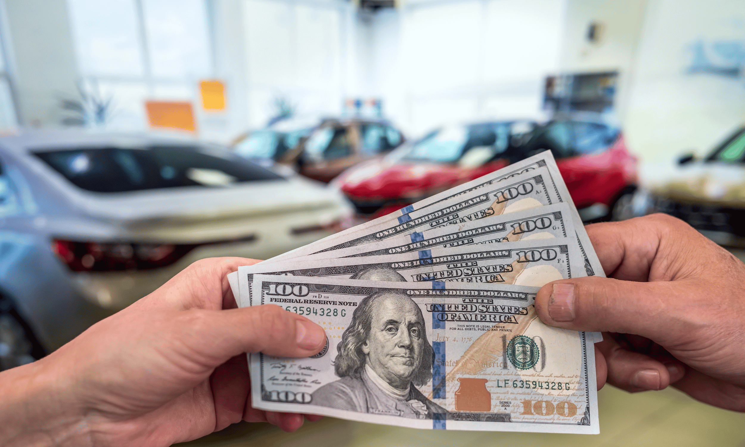 A person holding multiple $100 bills in front of a car dealership, with several cars inside the showroom.