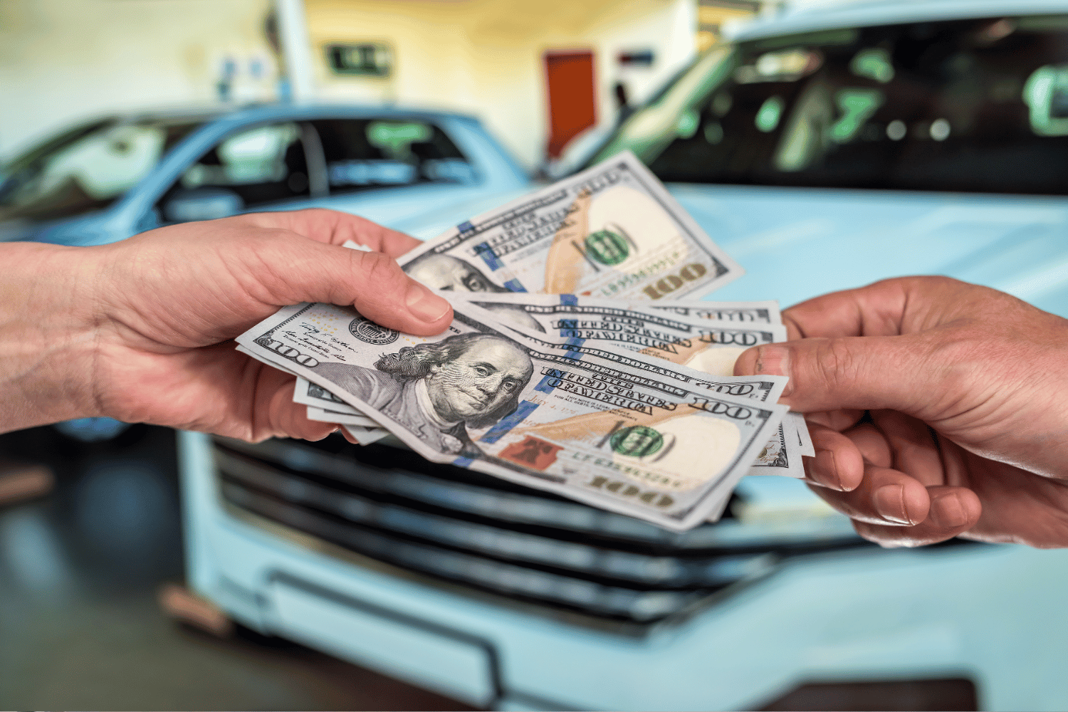 Two hands exchanging hundred dollar bills in front of a light blue car.