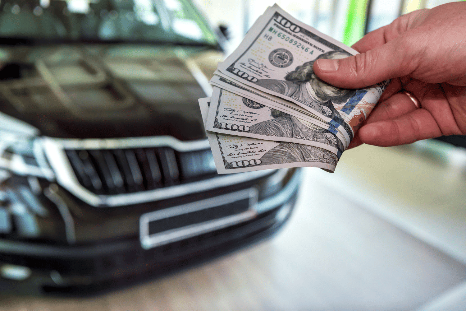 Hand holding several $100 bills in front of a black car.