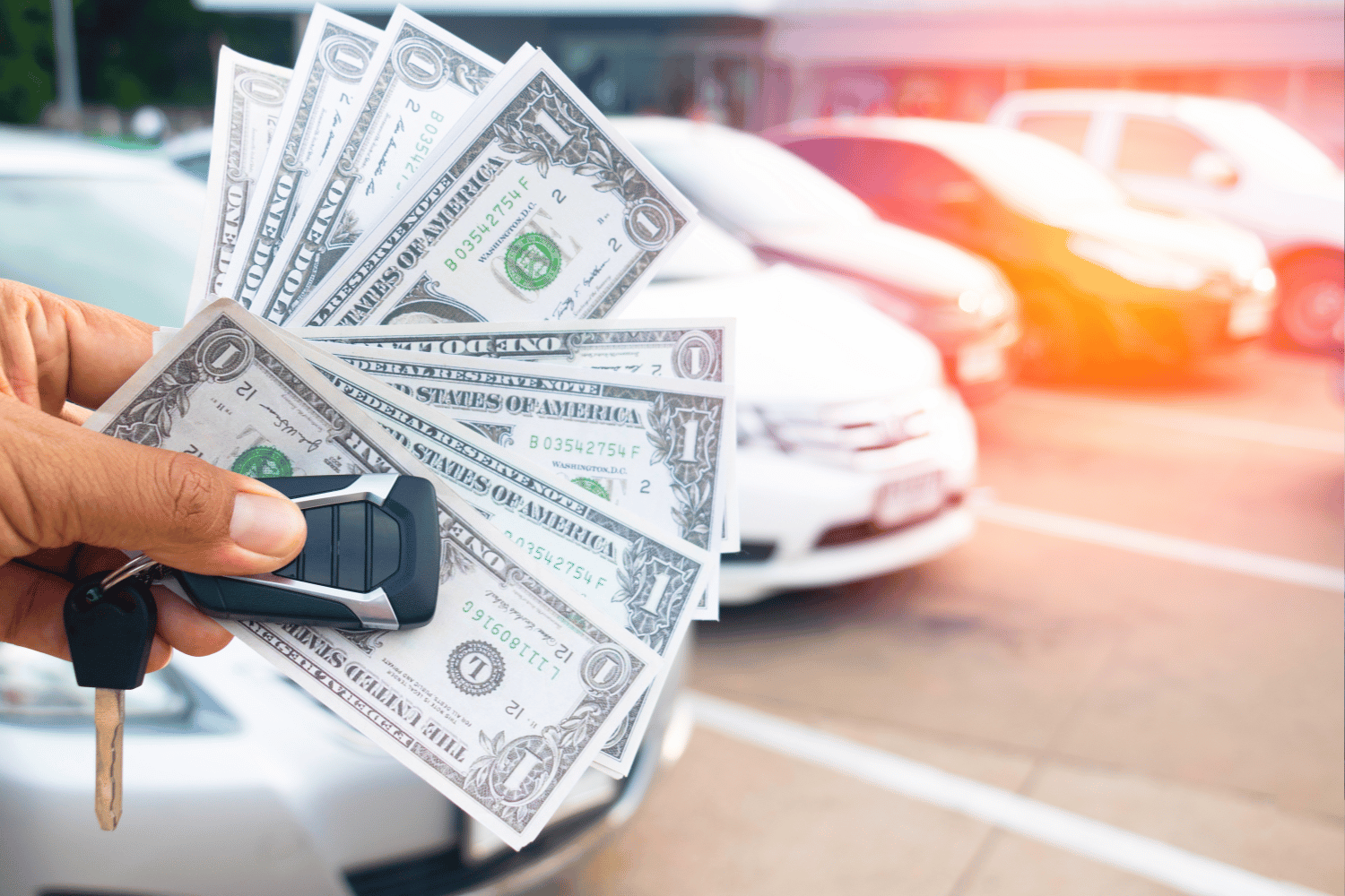 A hand holding a car key and several US dollar bills in front of parked cars in a parking lot.