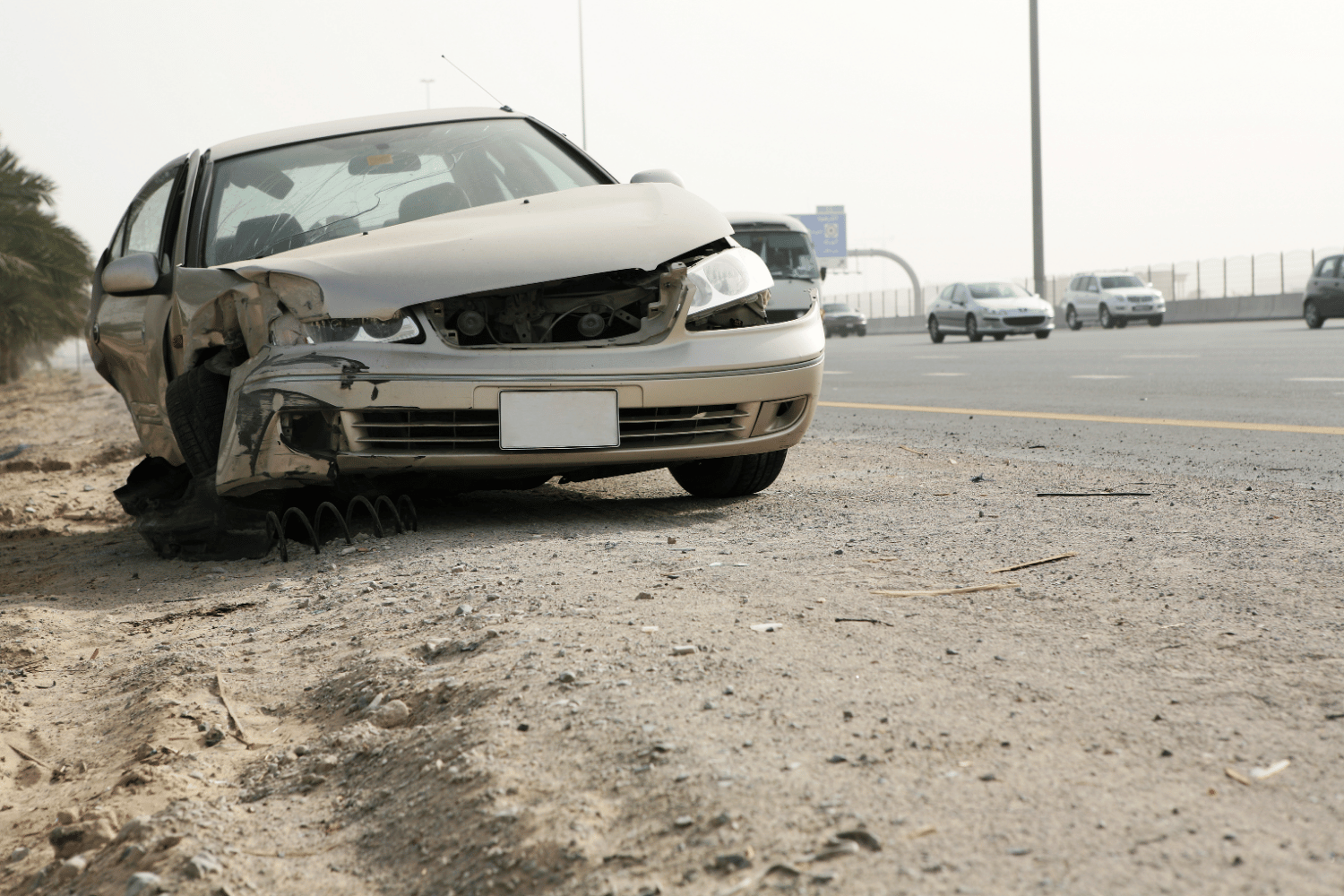 Damaged front of a white sedan car crashed on the side of a highway, with debris and broken parts on the road.