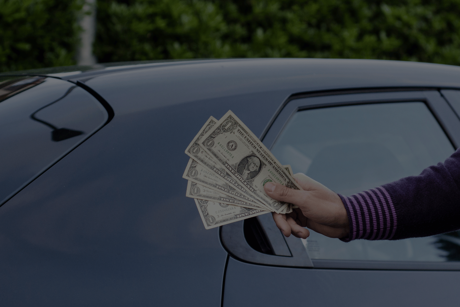 Hand holding multiple one-dollar bills being handed to a person through the car window.