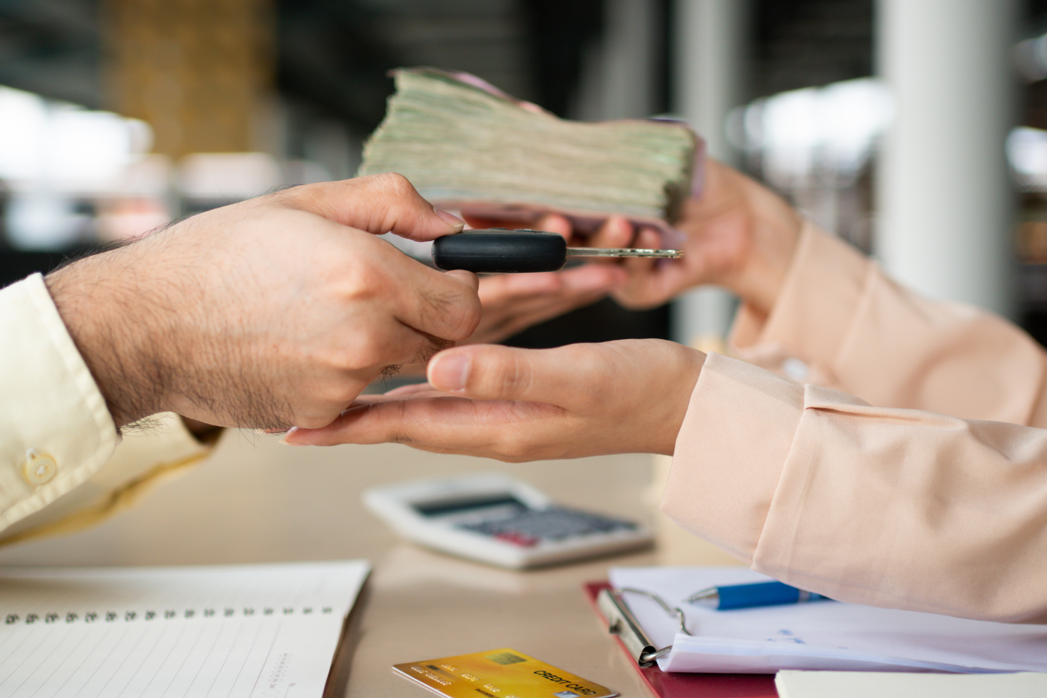 Person handing over keys and money to another person at a desk with notebooks, a calculator, and a credit card.