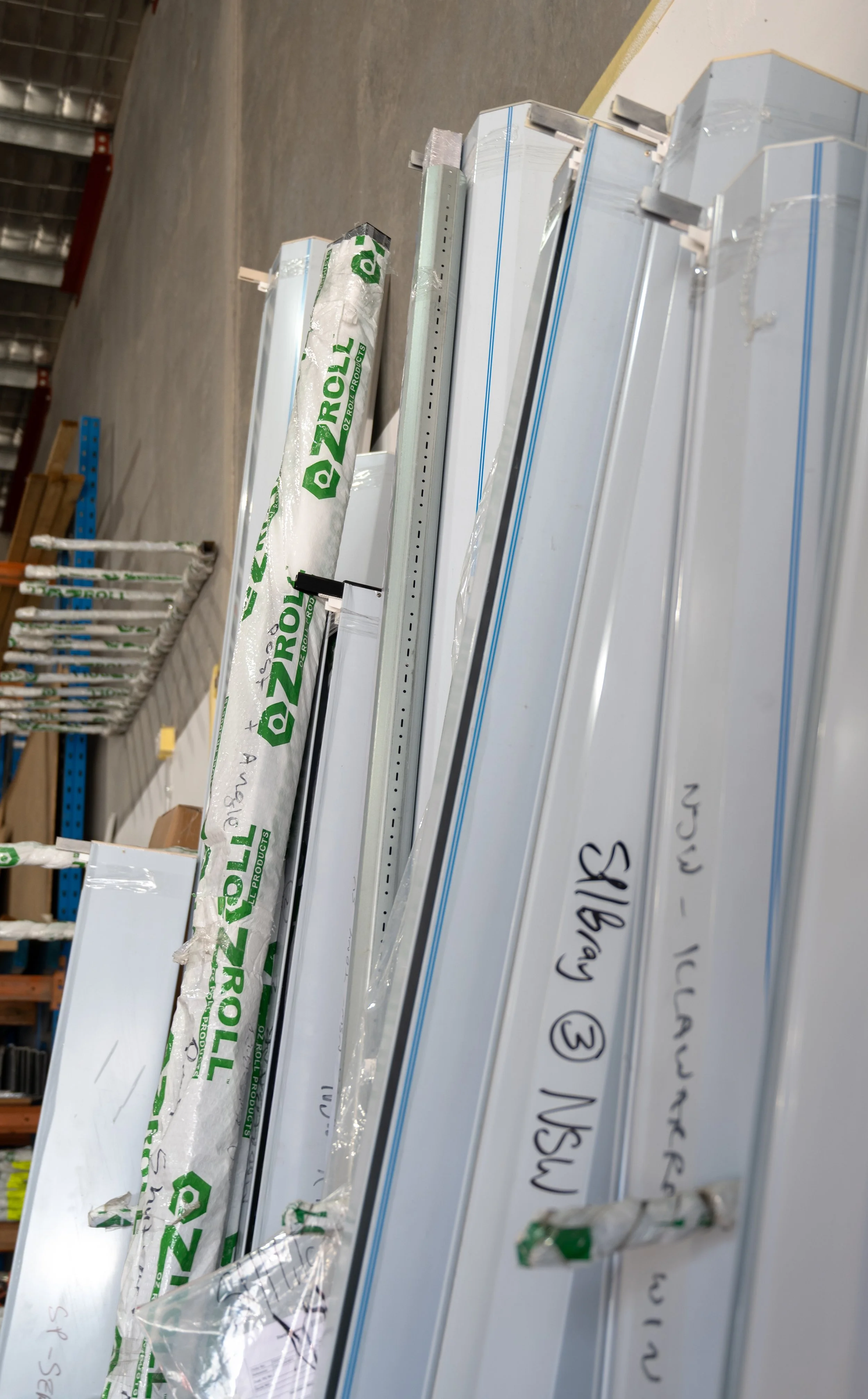 Stacked Roller Shutters in a warehouse, some with packaging wrapped around them, with handwritten labels and tags.